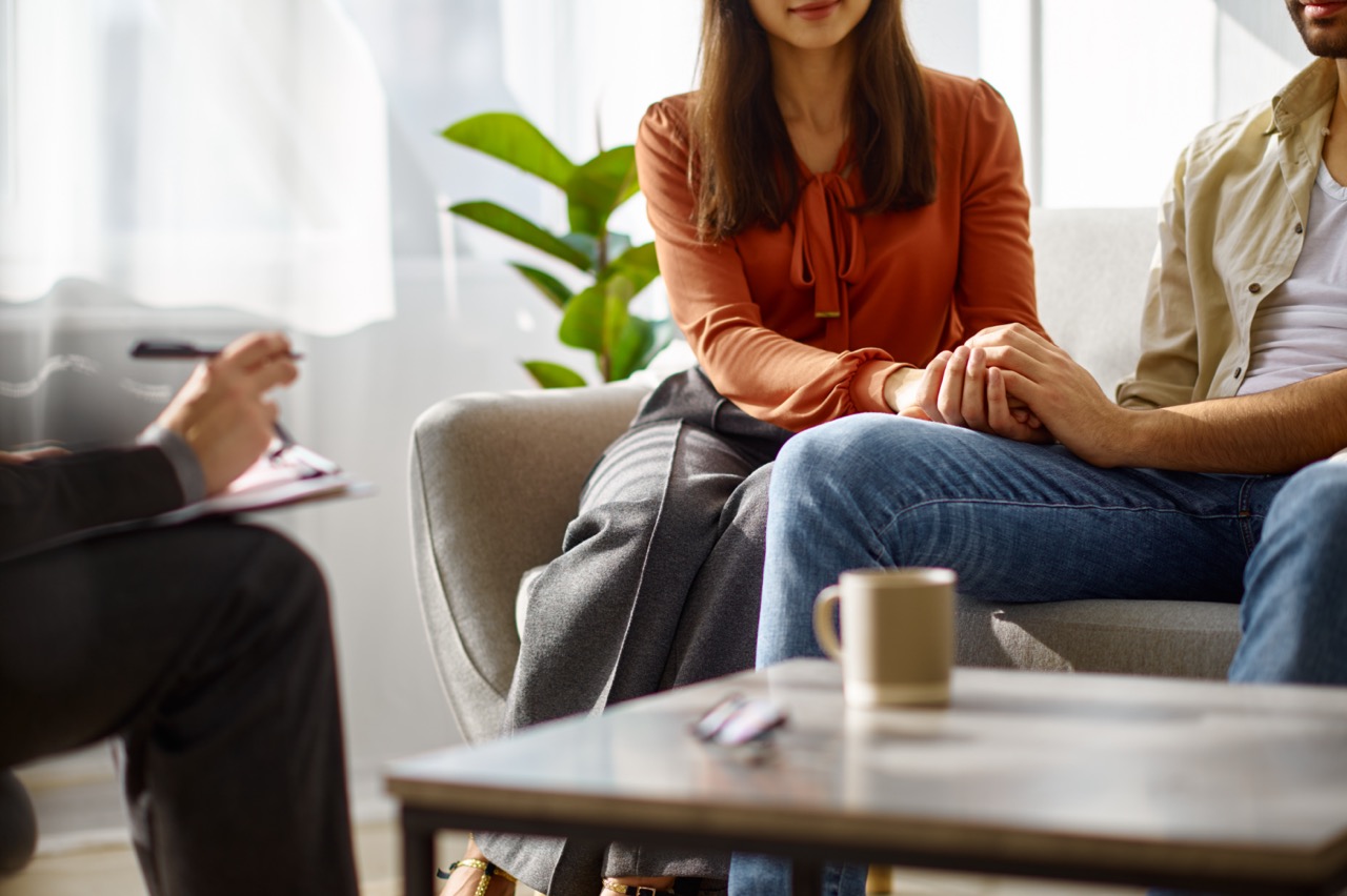 A couple sitting side by side on a couch holding hands, facing a counselor who is taking notes with a pen on a clipboard; a coffee mug and eyeglasses are on the table in front of them.