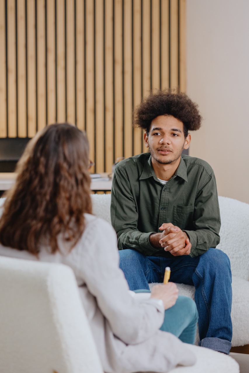 Young man with curly hair wearing a green shirt and jeans sitting on a white couch talking to a woman with long brown hair, who is holding a pen and notepad in a bright room with wooden paneling.