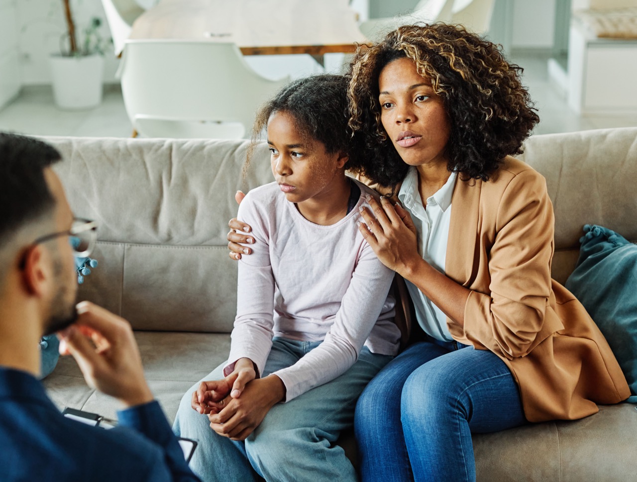 A concerned mother comforting her distressed daughter while sitting on a couch, engaged in a serious conversation with a man, likely a counselor, seated across from them.