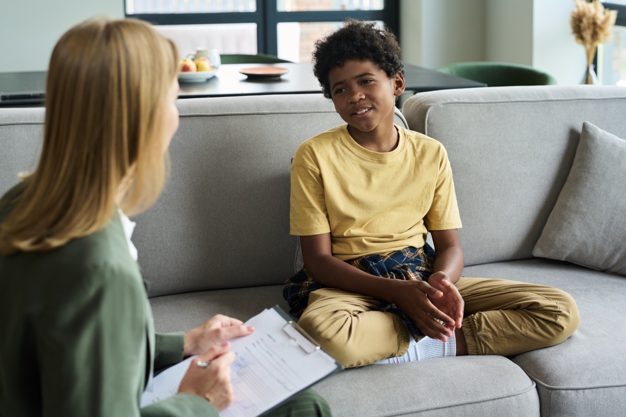 Young boy sitting cross-legged on a gray couch, engaged in conversation with a therapist holding a clipboard in a bright room with large windows.
