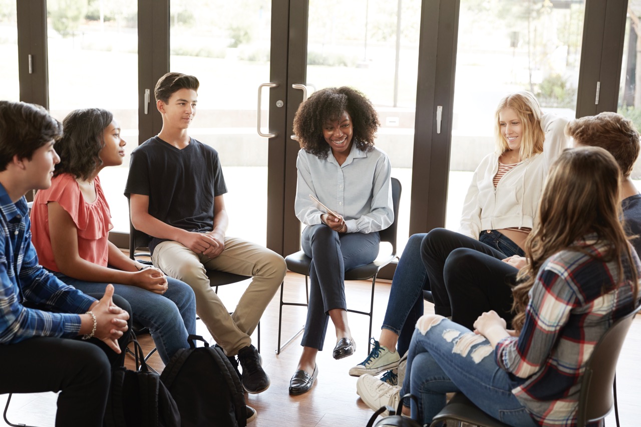 A female tutor leading a discussion in a casual group setting with six teenagers seated in a circle in a bright room with large glass doors.