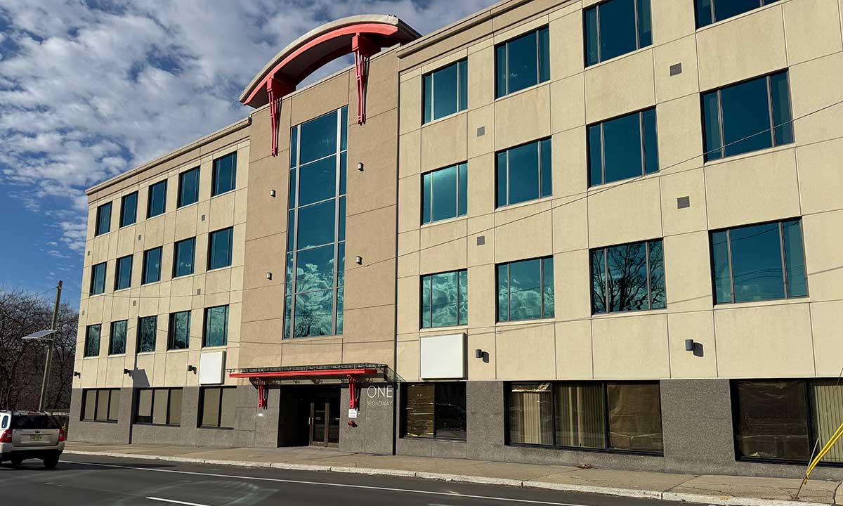 Three-story beige commercial building with large blue-tinted windows and red architectural accents at the entrance and rooftop, under a partly cloudy sky.