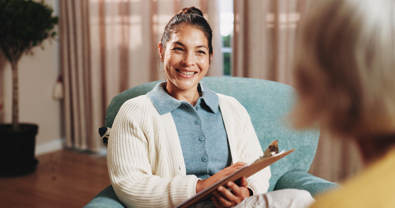 Smiling woman sitting in a teal armchair holding a clipboard, engaged in conversation with another person.
