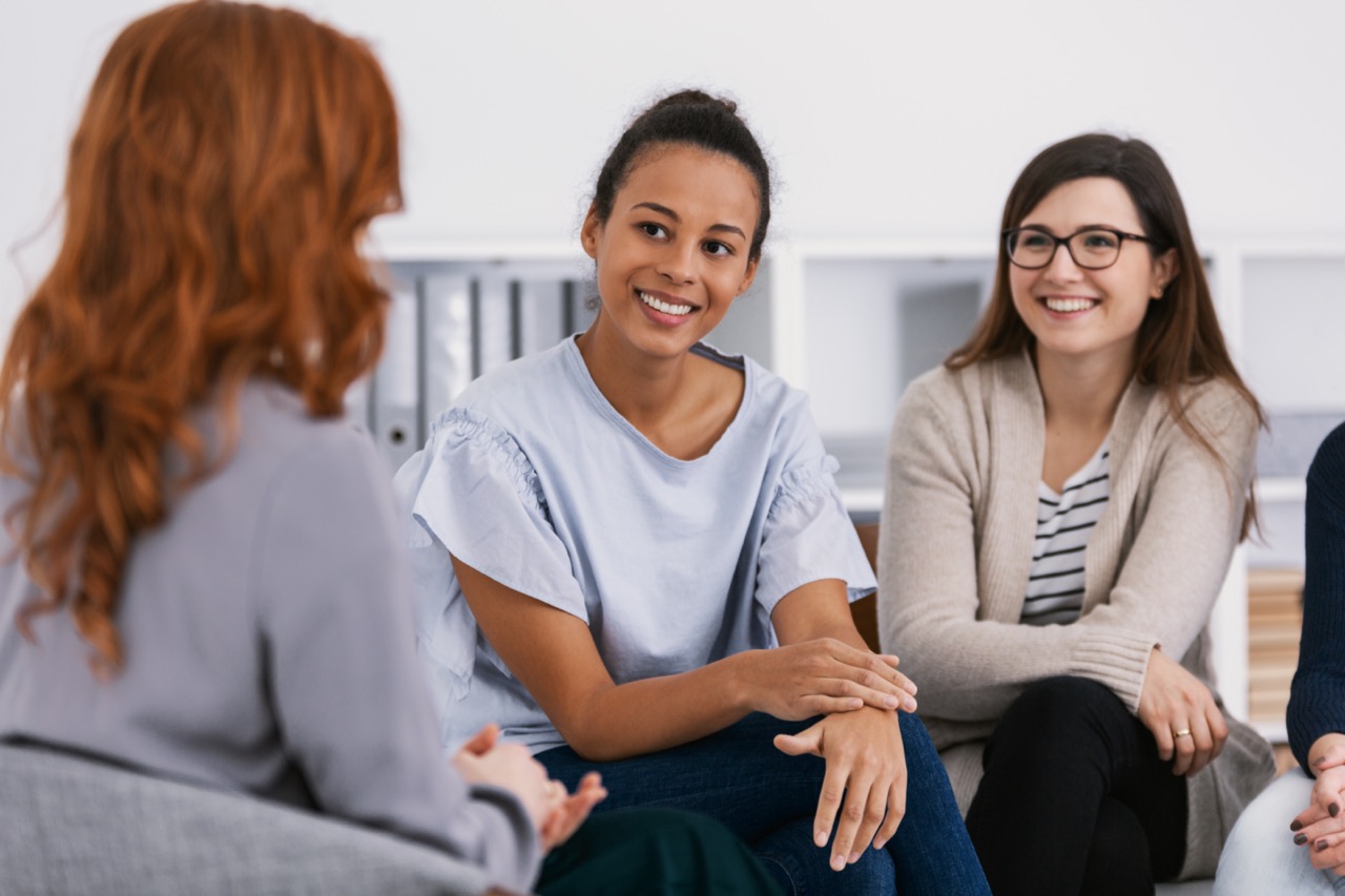 Three women sitting in a circle, engaged in a friendly and supportive conversation, smiling and listening attentively in a bright room with shelves in the background.