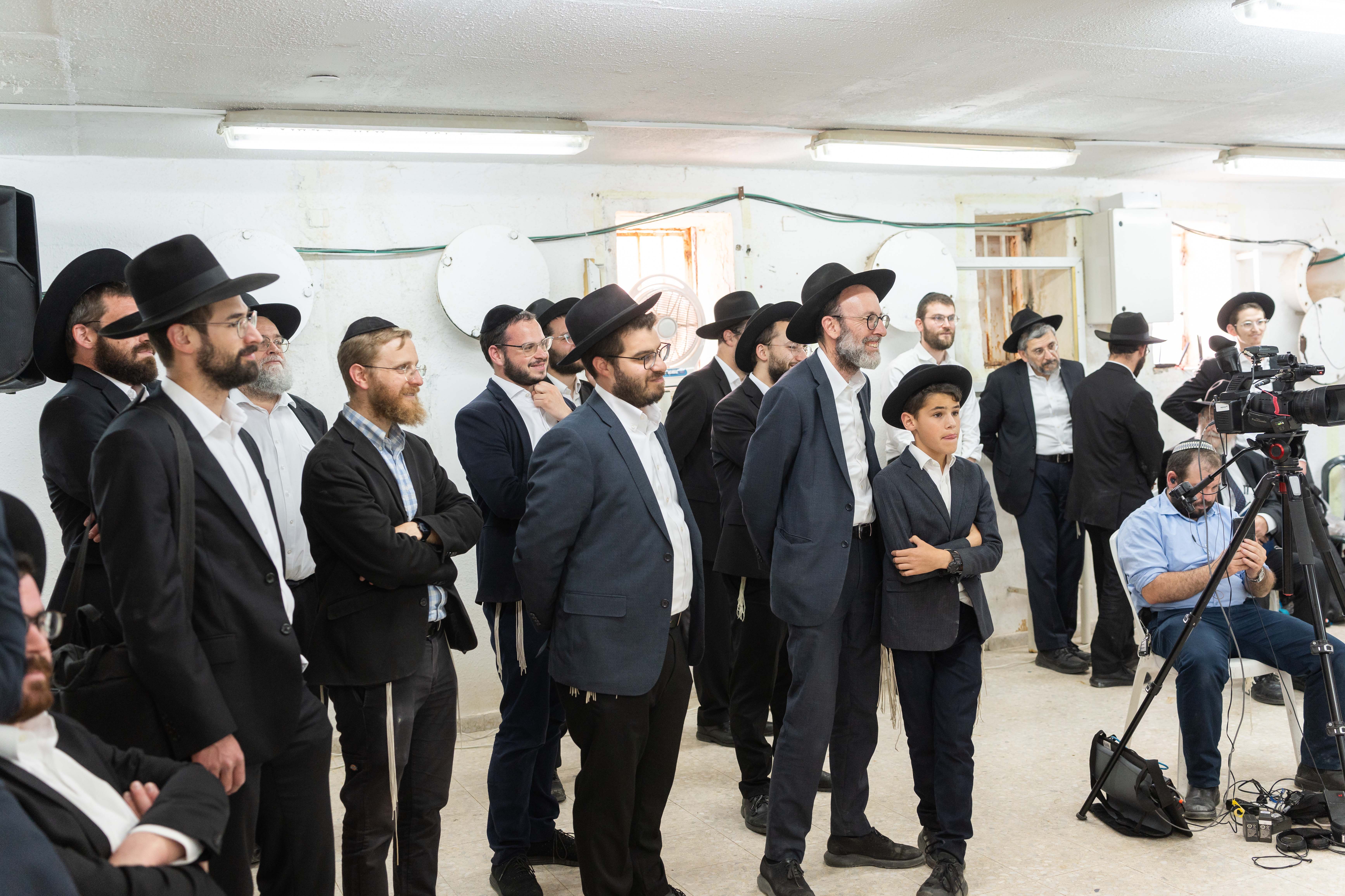 Group of men and boys in black hats and suits gathered in a room with camera equipment.