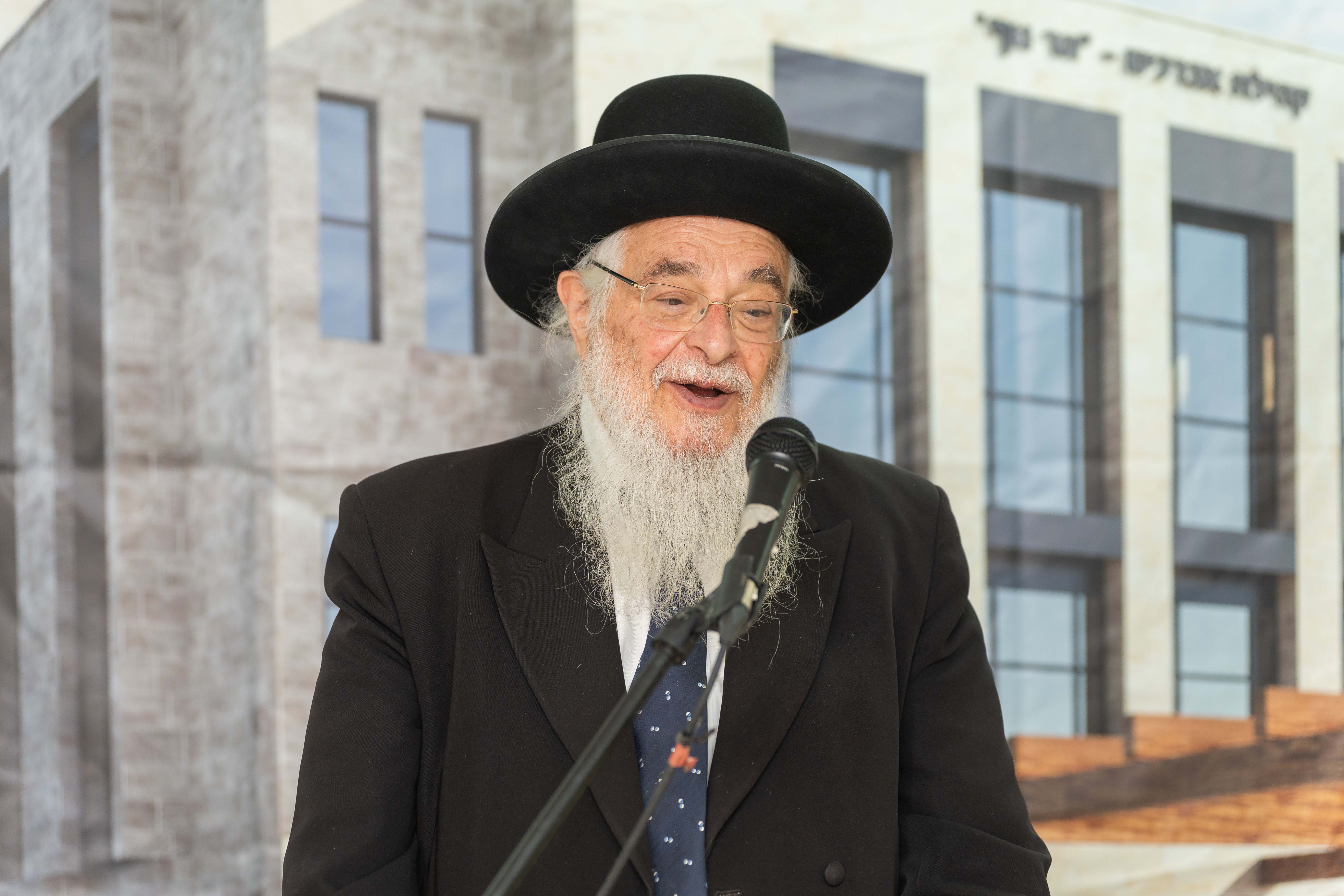 Elderly man with white beard and glasses speaking into a microphone outdoors in front of large building.