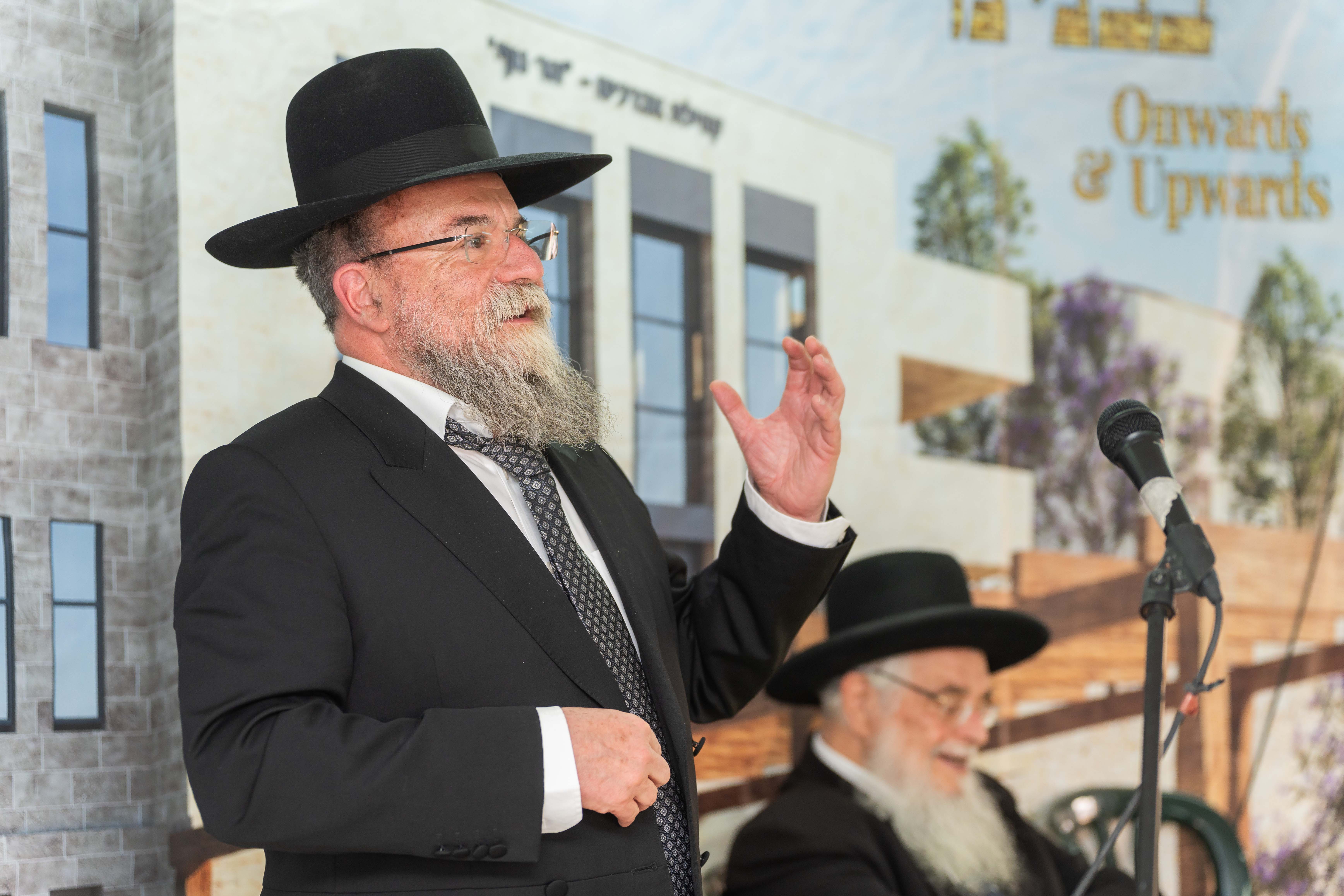 Bearded man in black suit and hat speaking, with another seated man and microphone in background.