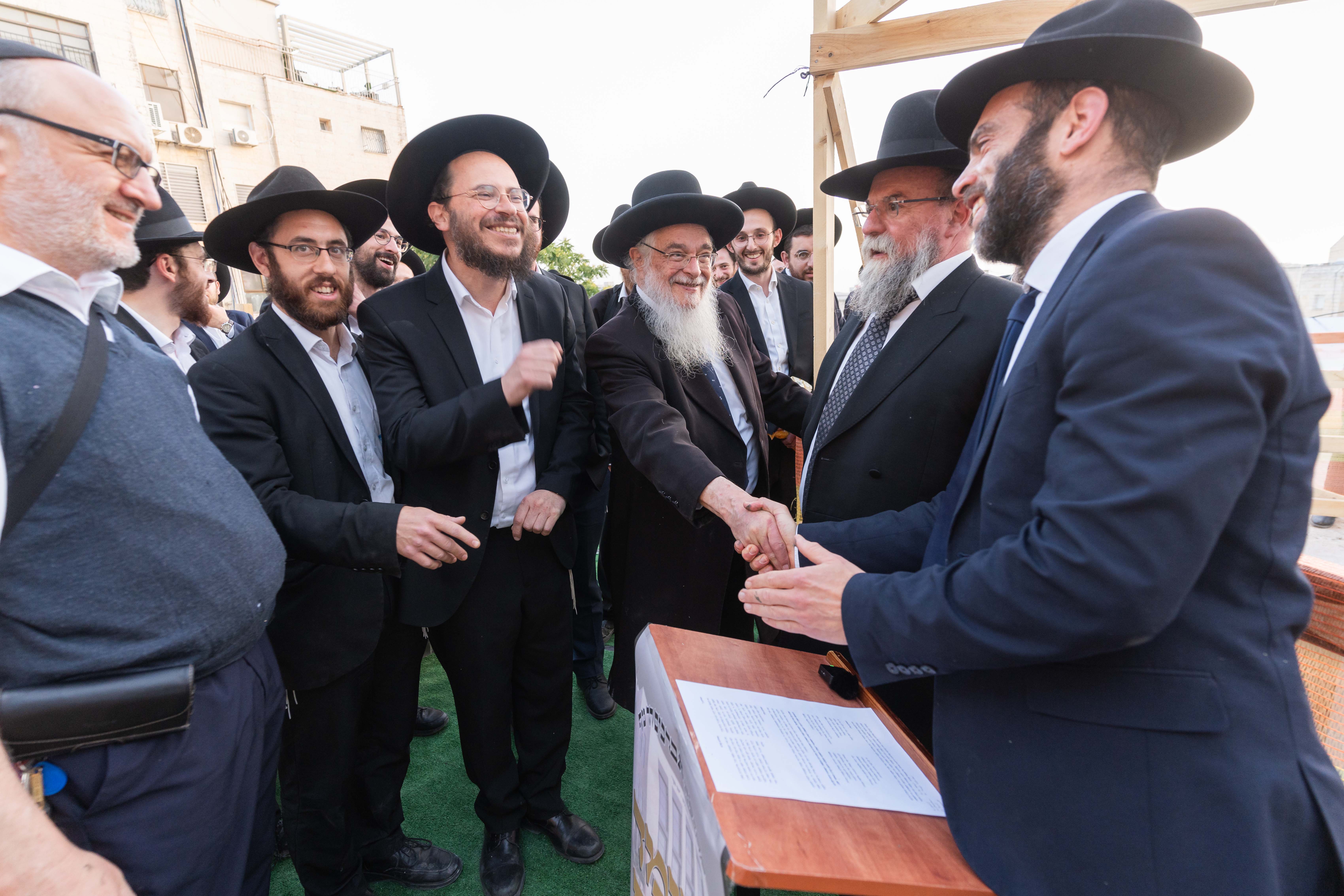 Group of men in black hats and suits smiling and shaking hands outdoors on green carpet.