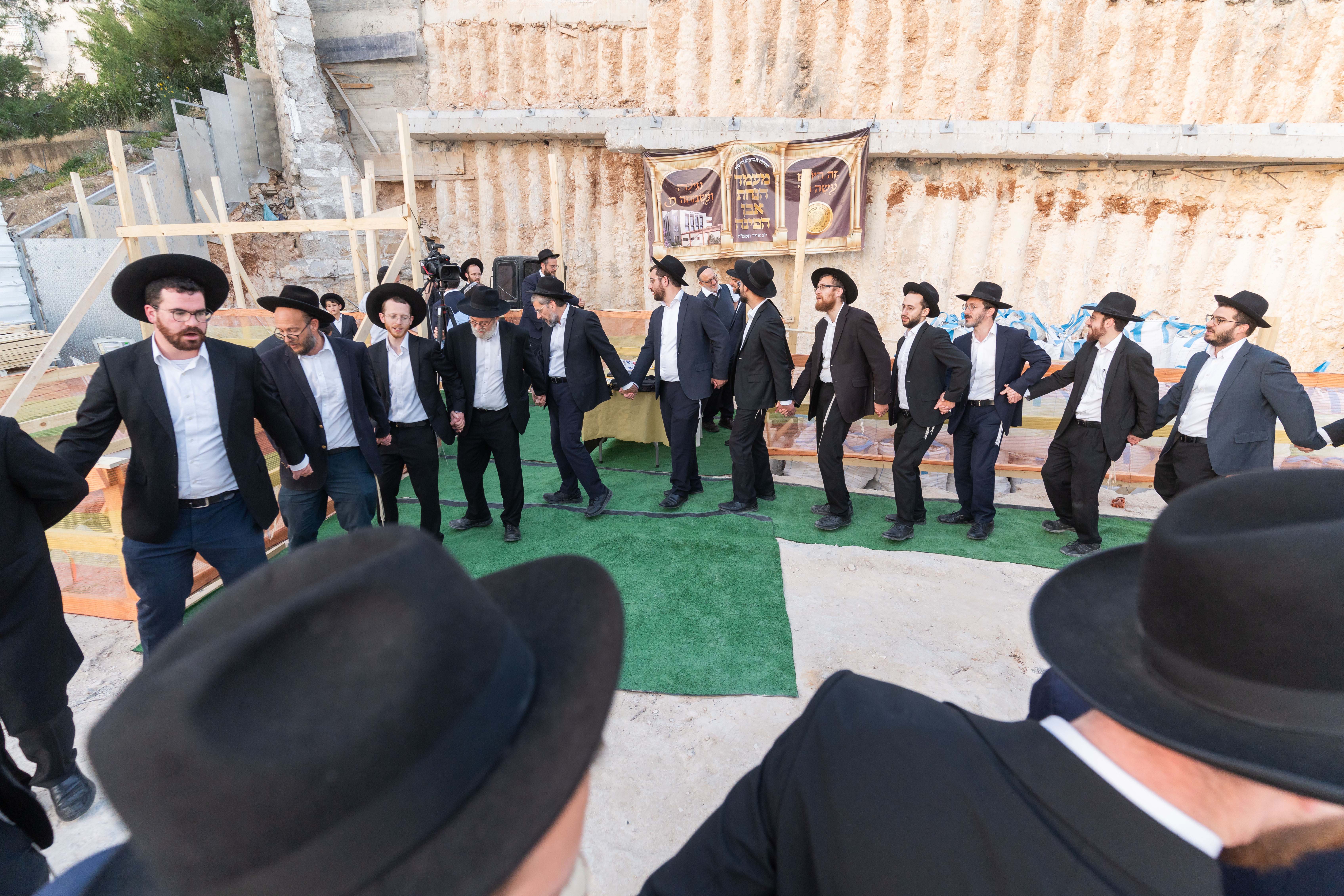 Group of men in black hats and suits holding hands in a circle outdoors near a stone wall.