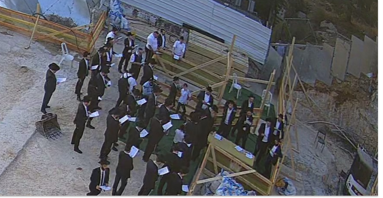 Group of men in black suits holding papers, standing in and around wooden construction frames outdoors.