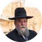 Bearded man wearing glasses and a wide-brimmed black hat in front of a stone wall.