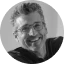 Smiling man with glasses and gray hair wearing a dark patterned shirt and dark jacket.