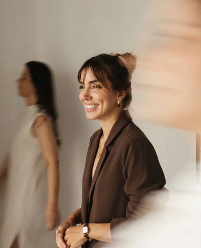 Smiling woman with a brown blazer and jewelry standing indoors with blurred people in background.