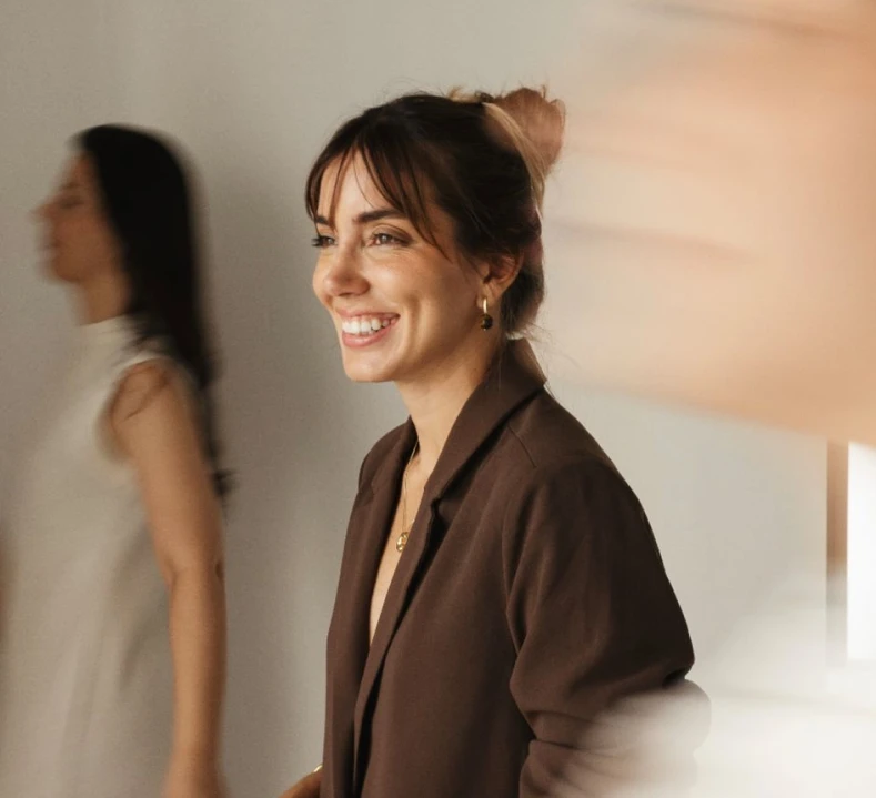 Smiling woman with a brown blazer and jewelry standing indoors with blurred people in background.