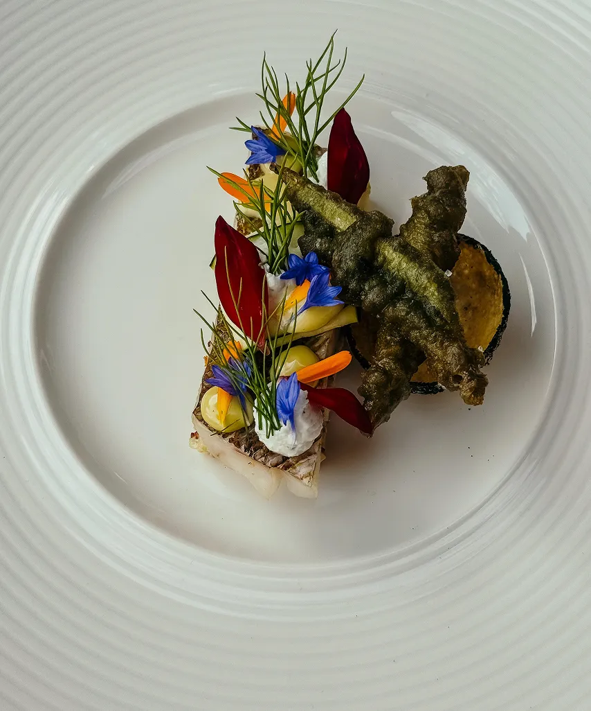 Gourmet dish with fish, edible flowers, herbs, and tempura vegetables on a white plate.