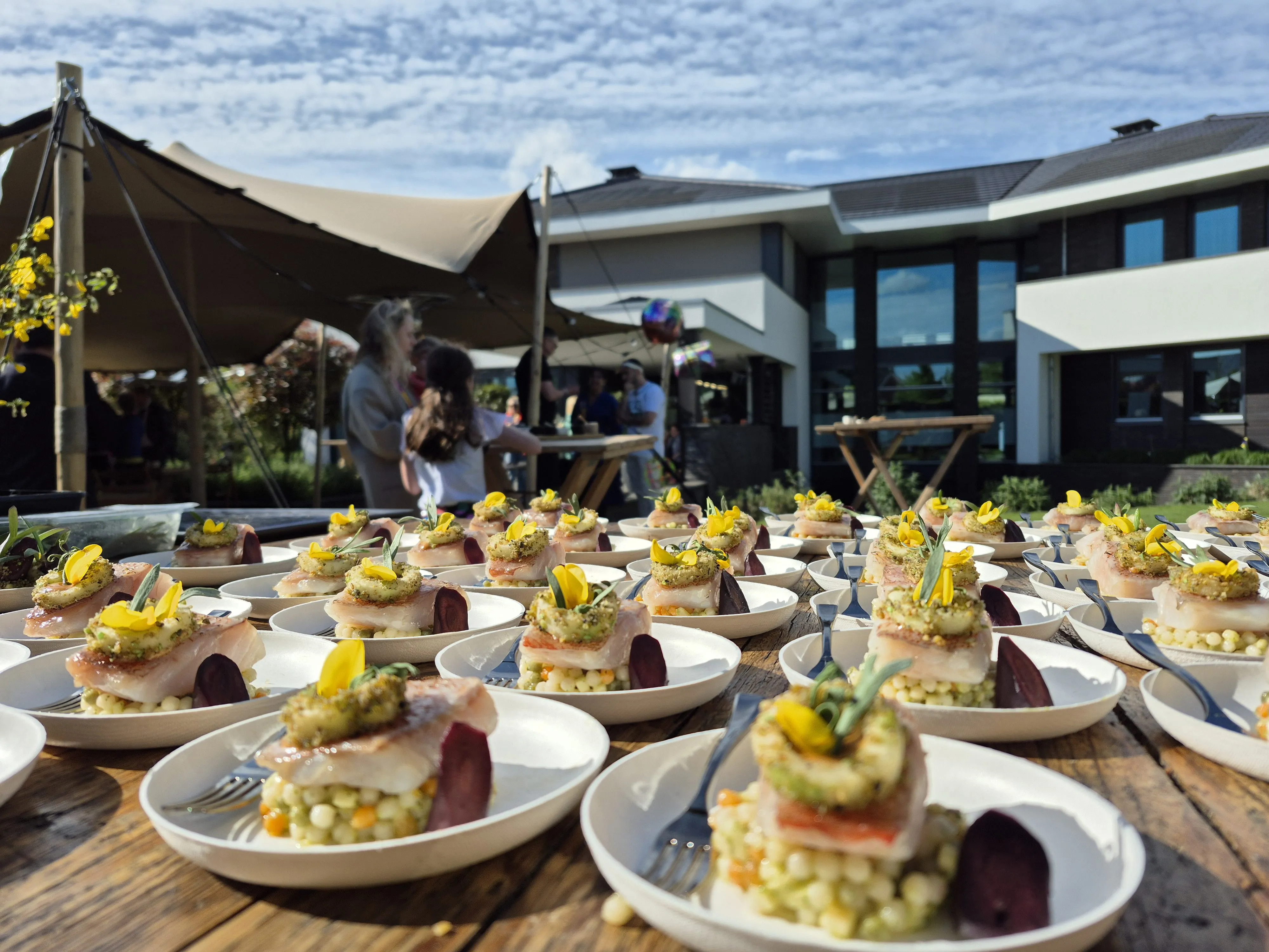 Plates of gourmet dishes with fish and yellow edible flowers on a wooden table outdoors.