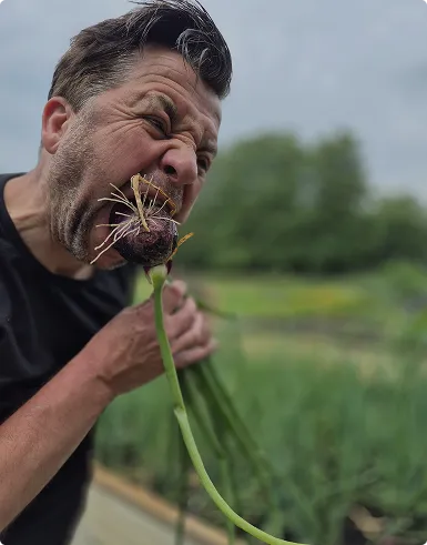 Man grimacing while biting the bulb of a green onion in a garden setting.