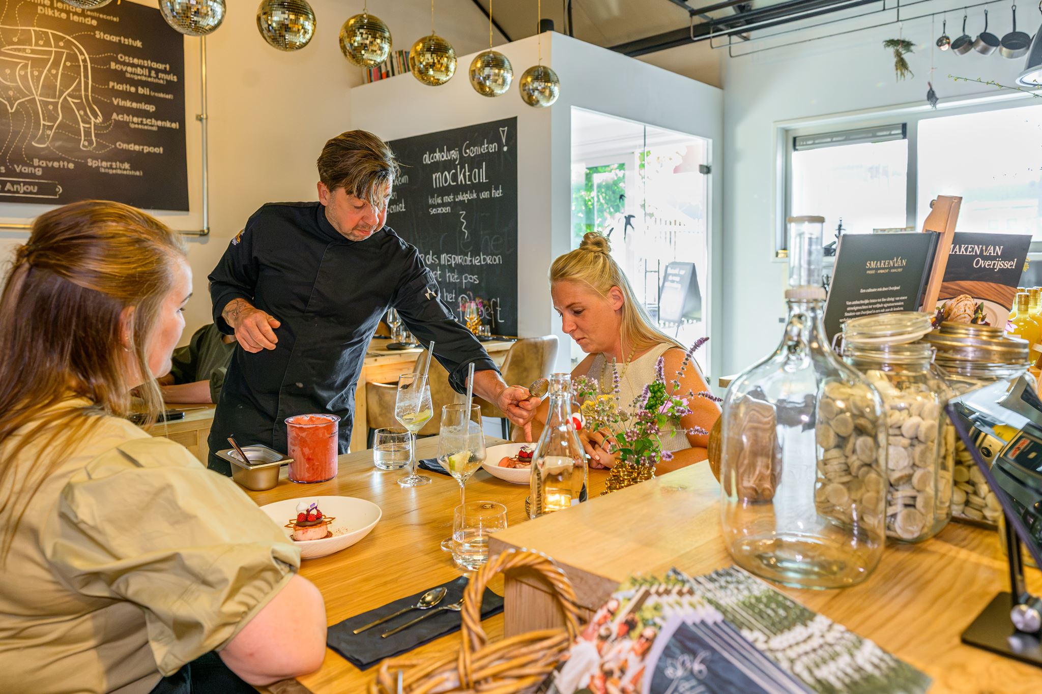Chef serving dessert to two women seated at a wooden counter in a casual restaurant.