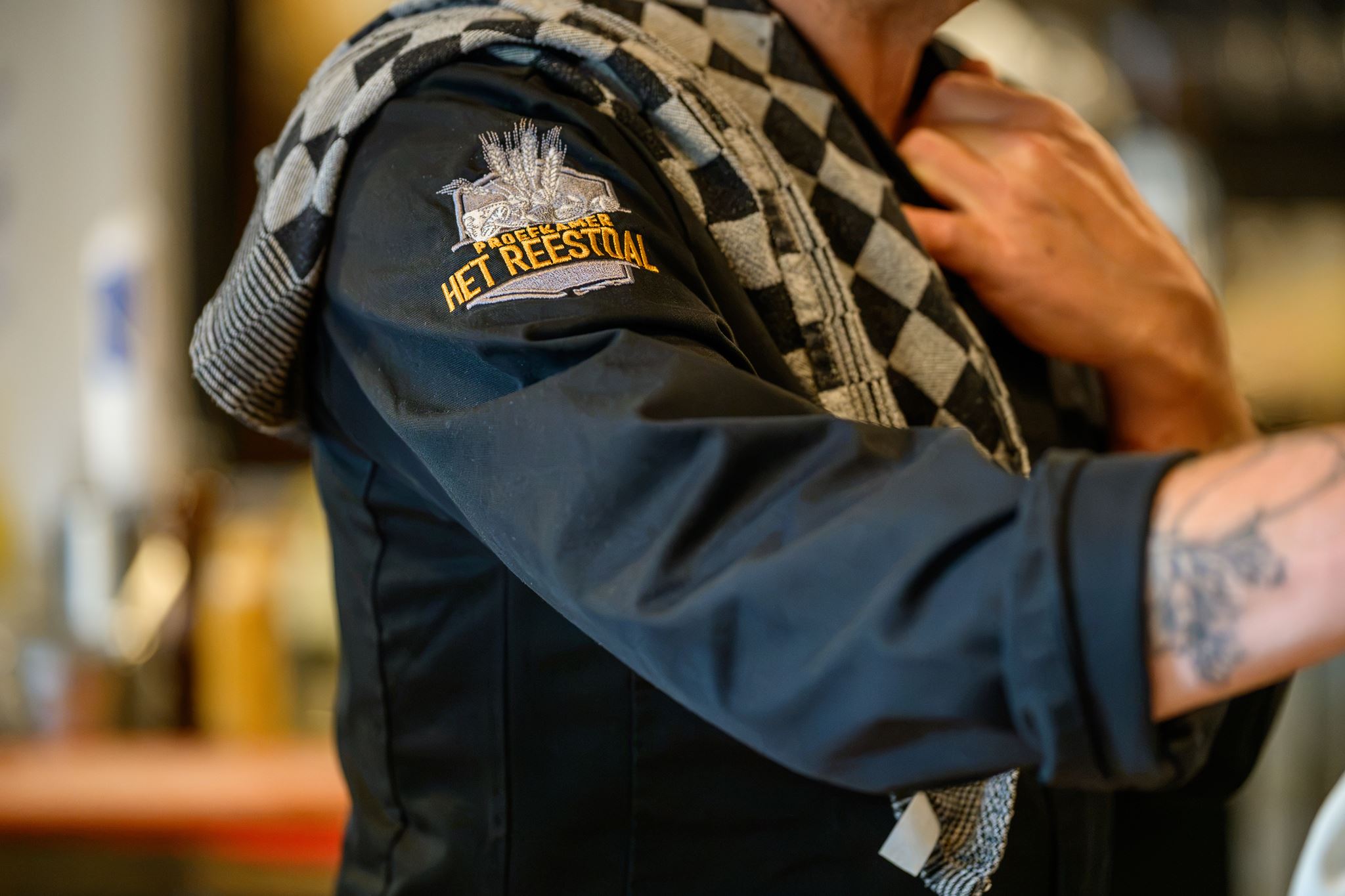 Person wearing a black chef jacket with logo and a checkered towel draped over their shoulder.