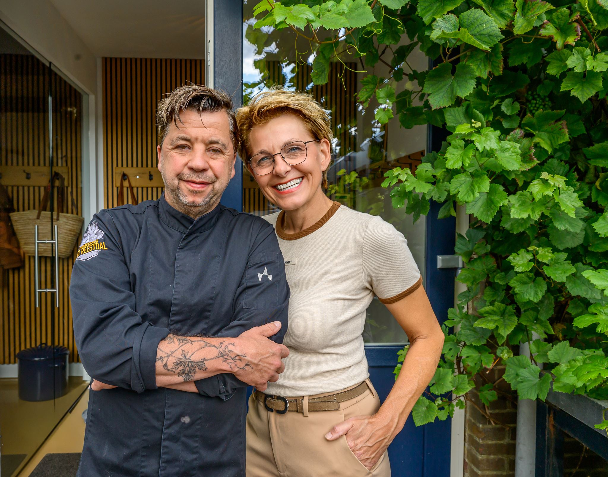 Smiling man in black chef coat with tattoo and woman in glasses and beige clothes standing outdoors by green leaves.