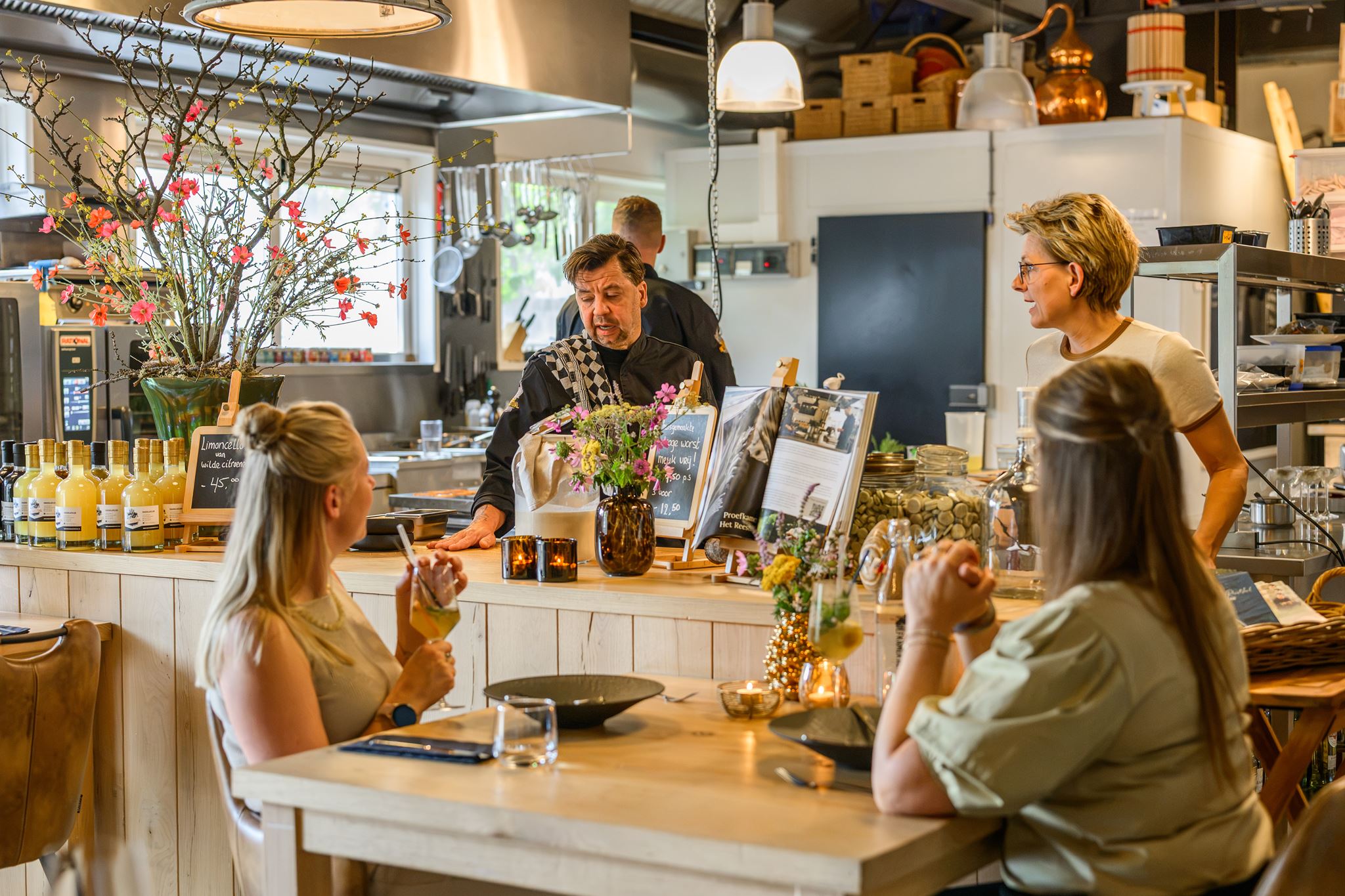 Two women seated at a table in a cozy restaurant, talking with two staff behind the counter.