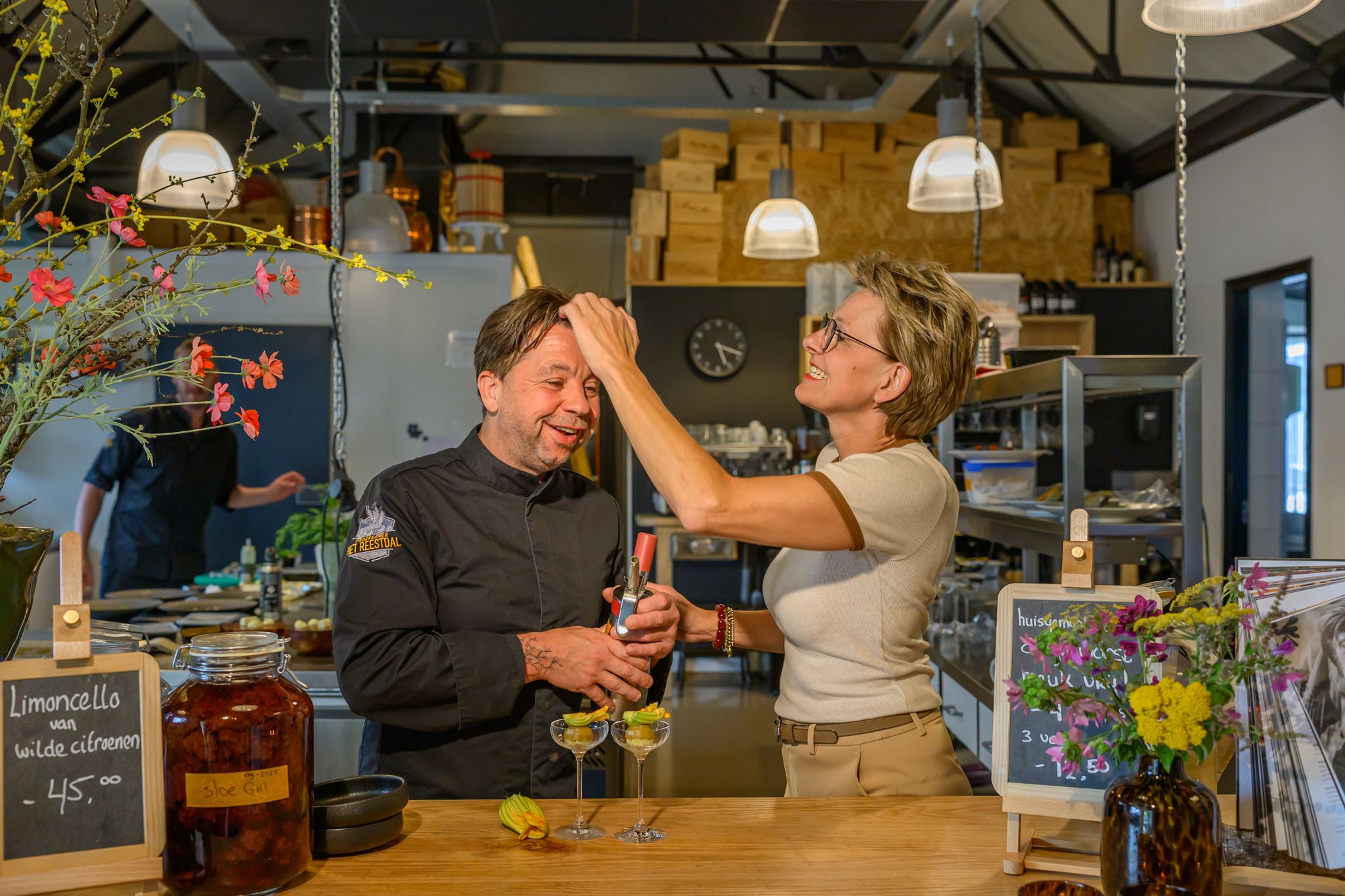 Woman playfully touching a male chef's hair while they laugh behind a bar counter with drinks and flowers.
