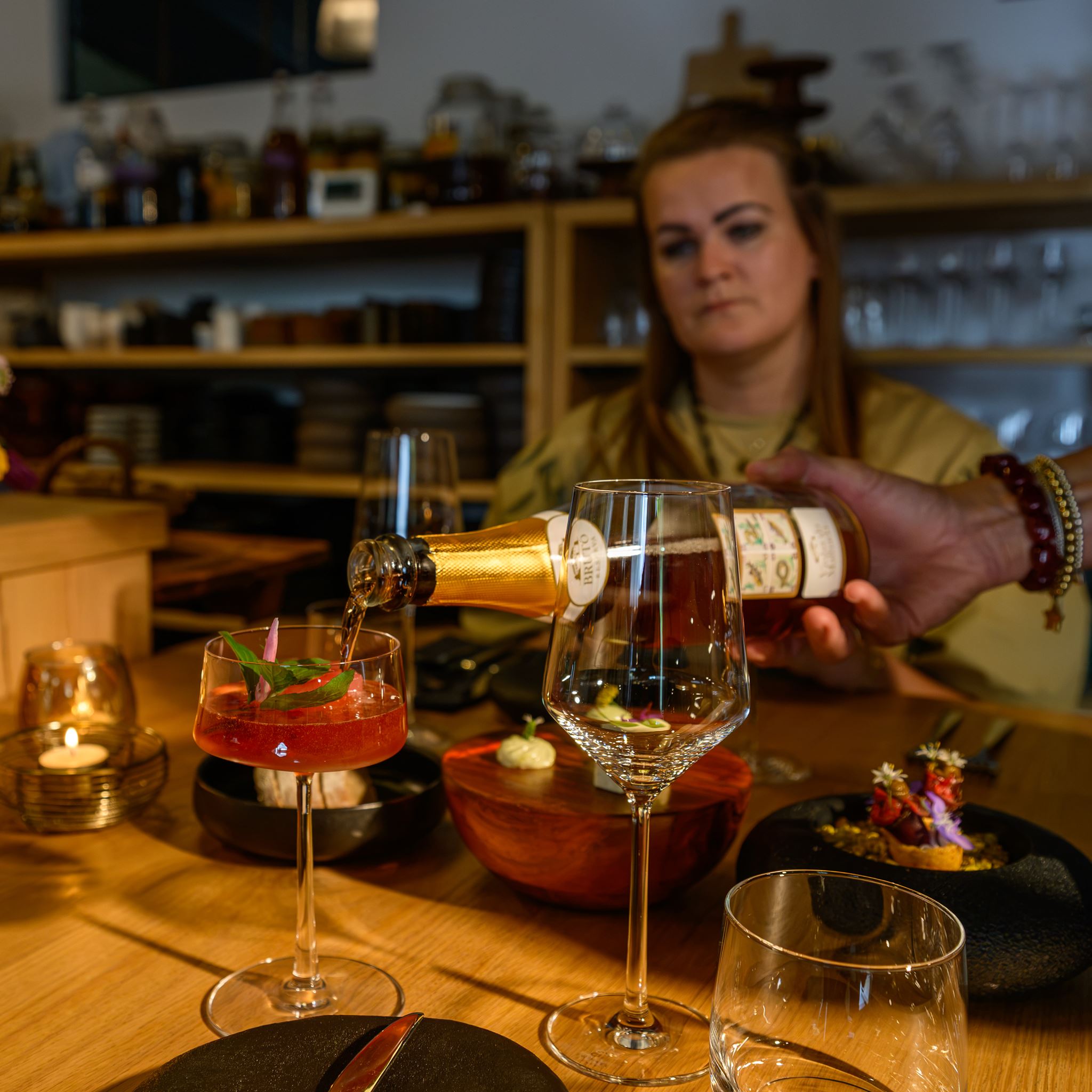 Hand pouring sparkling wine into a cocktail glass at a table with food and a person in the background.