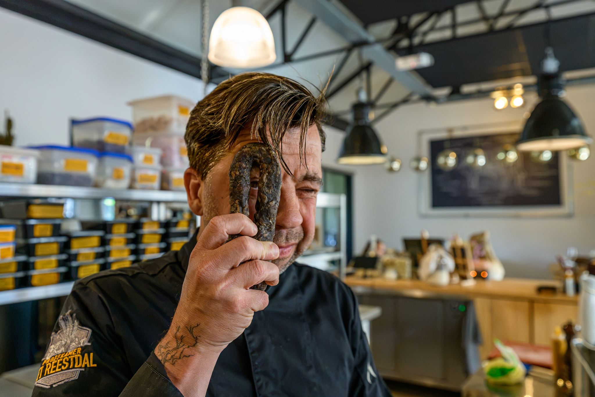 Man in black chef jacket holding a dark sausage loop up to his eye in a kitchen setting.