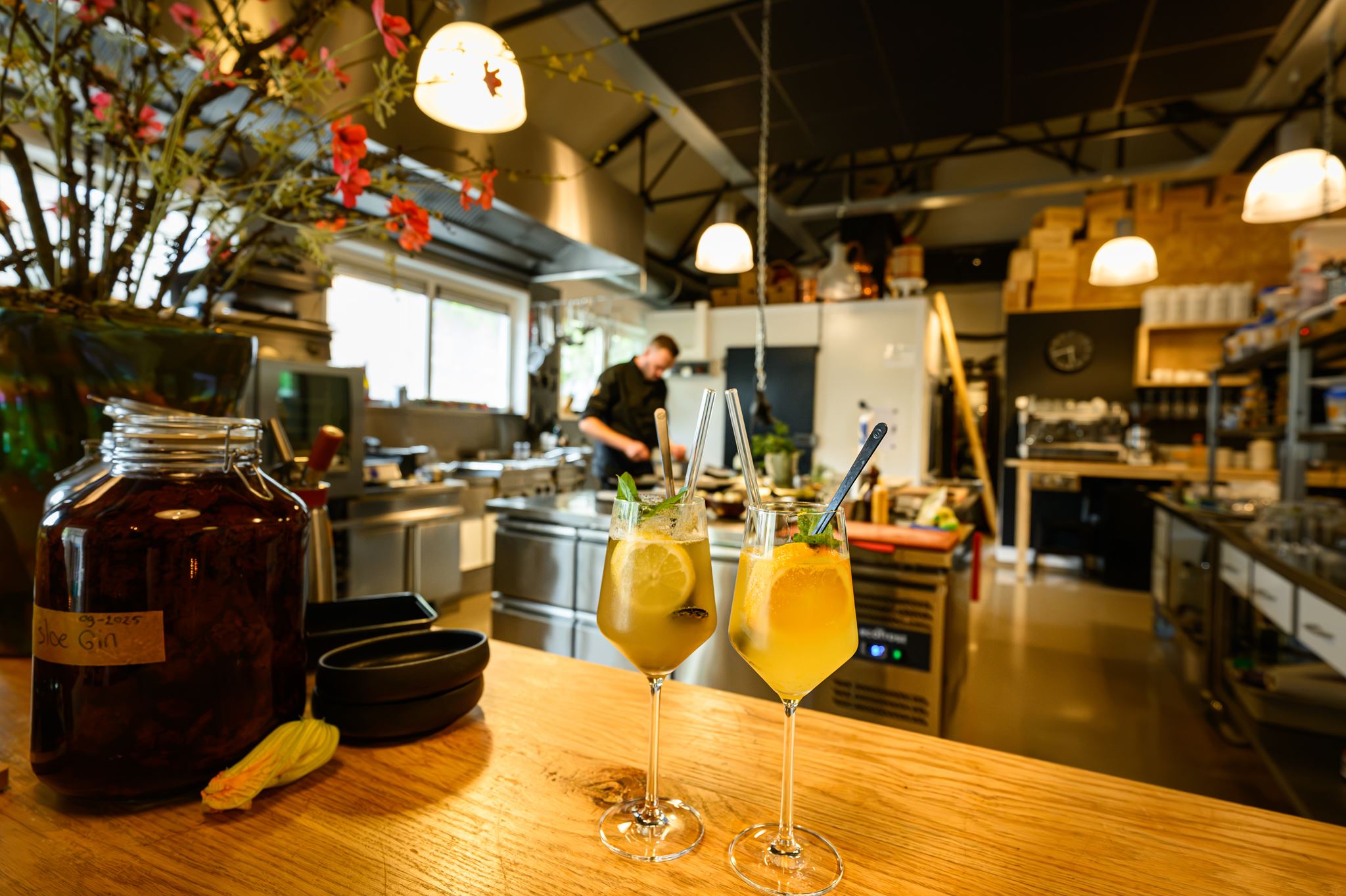 Two glasses with orange drinks and lemon slices on a wooden counter in a busy kitchen.