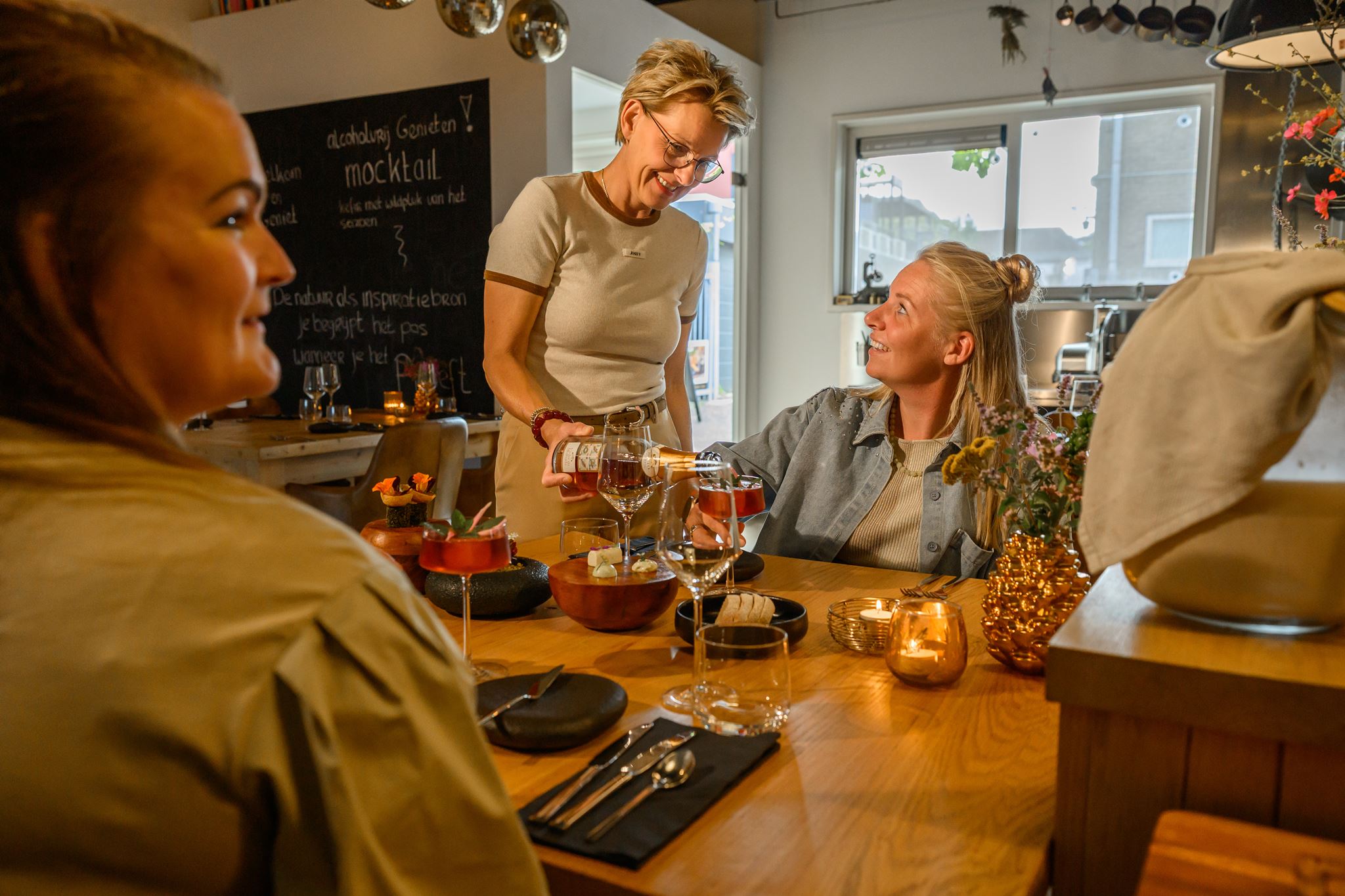 Woman pours wine into glass for seated woman at a warmly lit dining table with candles and food.