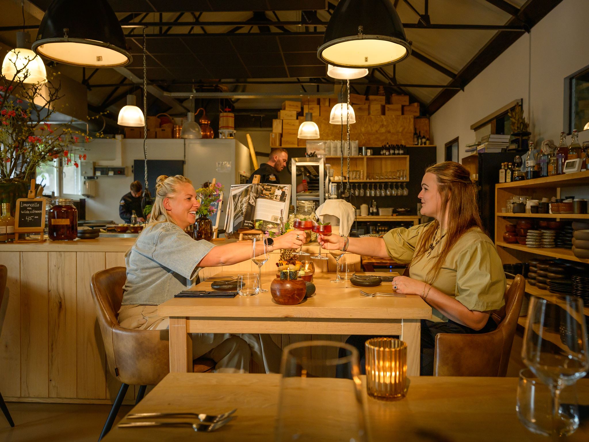 Two women sitting at a wooden table in a cozy restaurant, clinking glasses of red cocktail.