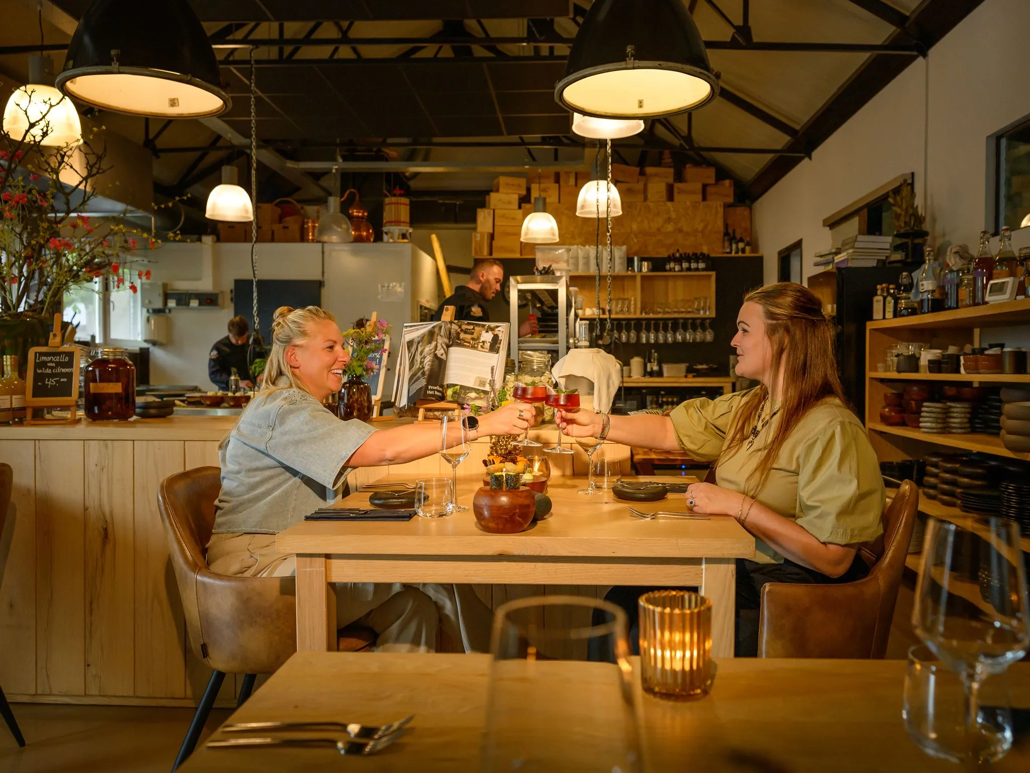 Two women sitting at a wooden table in a cozy restaurant, clinking glasses of red cocktail.