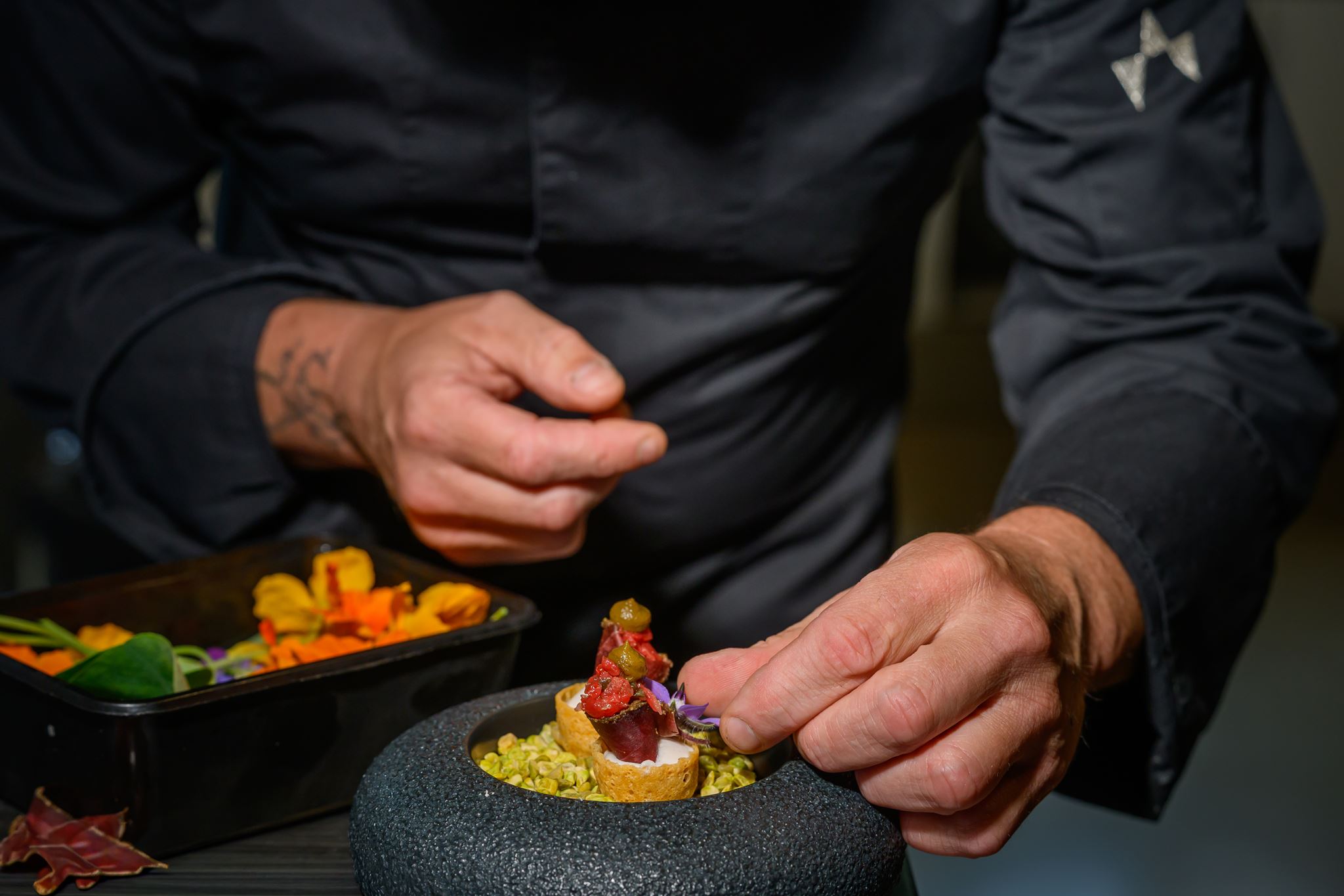 Chef garnishing intricate bite-sized appetizers with edible flowers on pistachios in a black bowl.