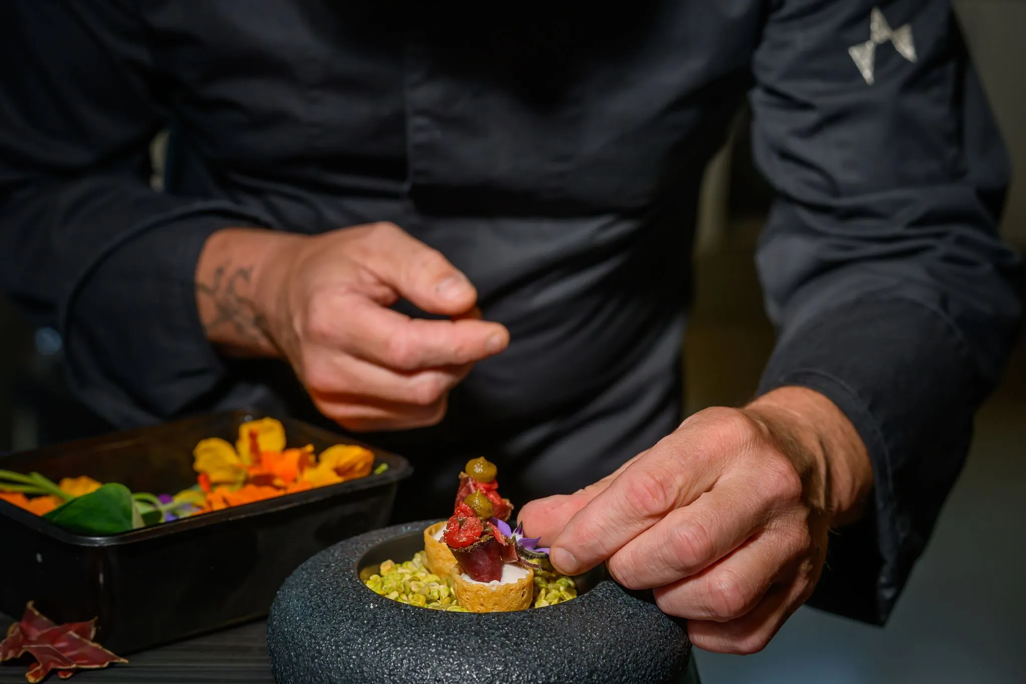Chef garnishing intricate bite-sized appetizers with edible flowers on pistachios in a black bowl.