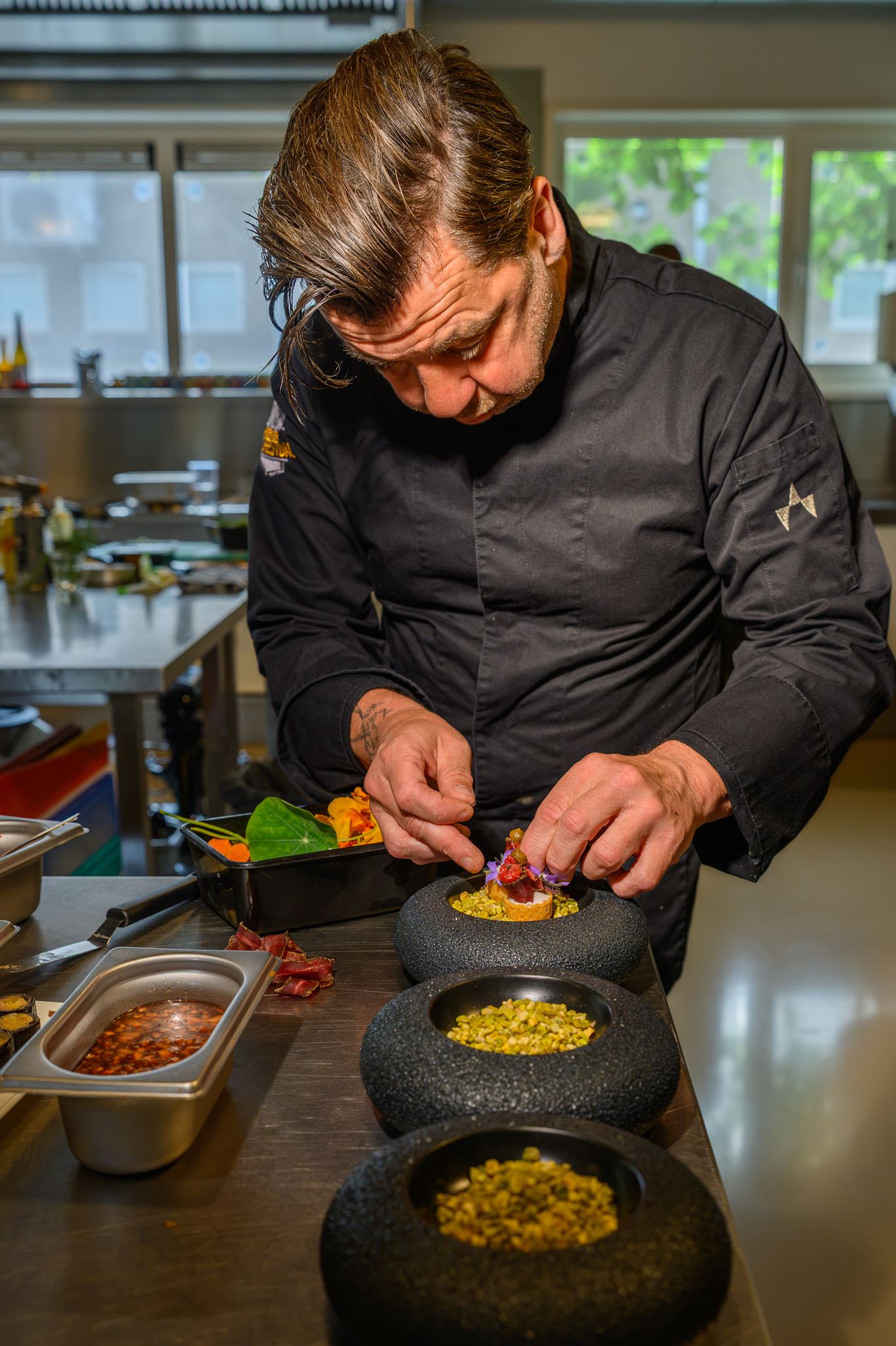 Chef in black uniform carefully placing edible flowers on gourmet appetizer in a professional kitchen.