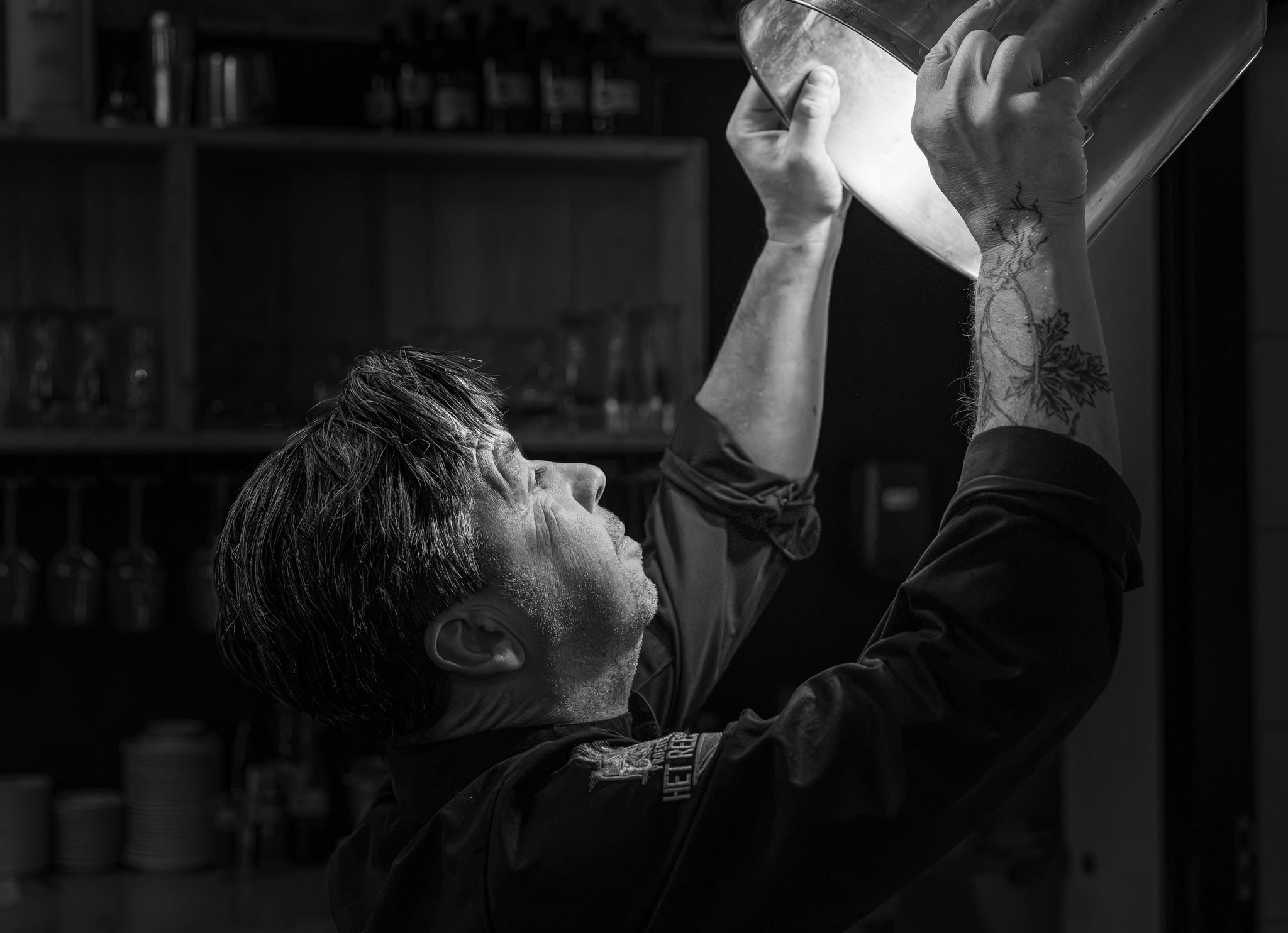 Man with tattooed arm holding a large metal bowl overhead inspecting its contents in a dim kitchen.