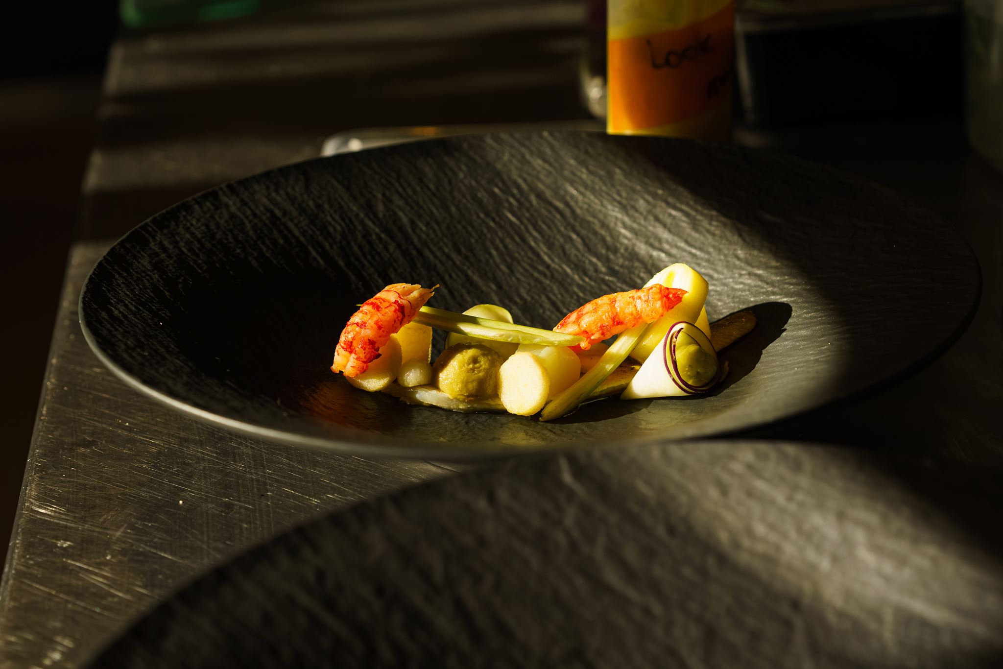 Gourmet dish with two shrimp, green sauce, and white vegetable slices on a black textured plate.