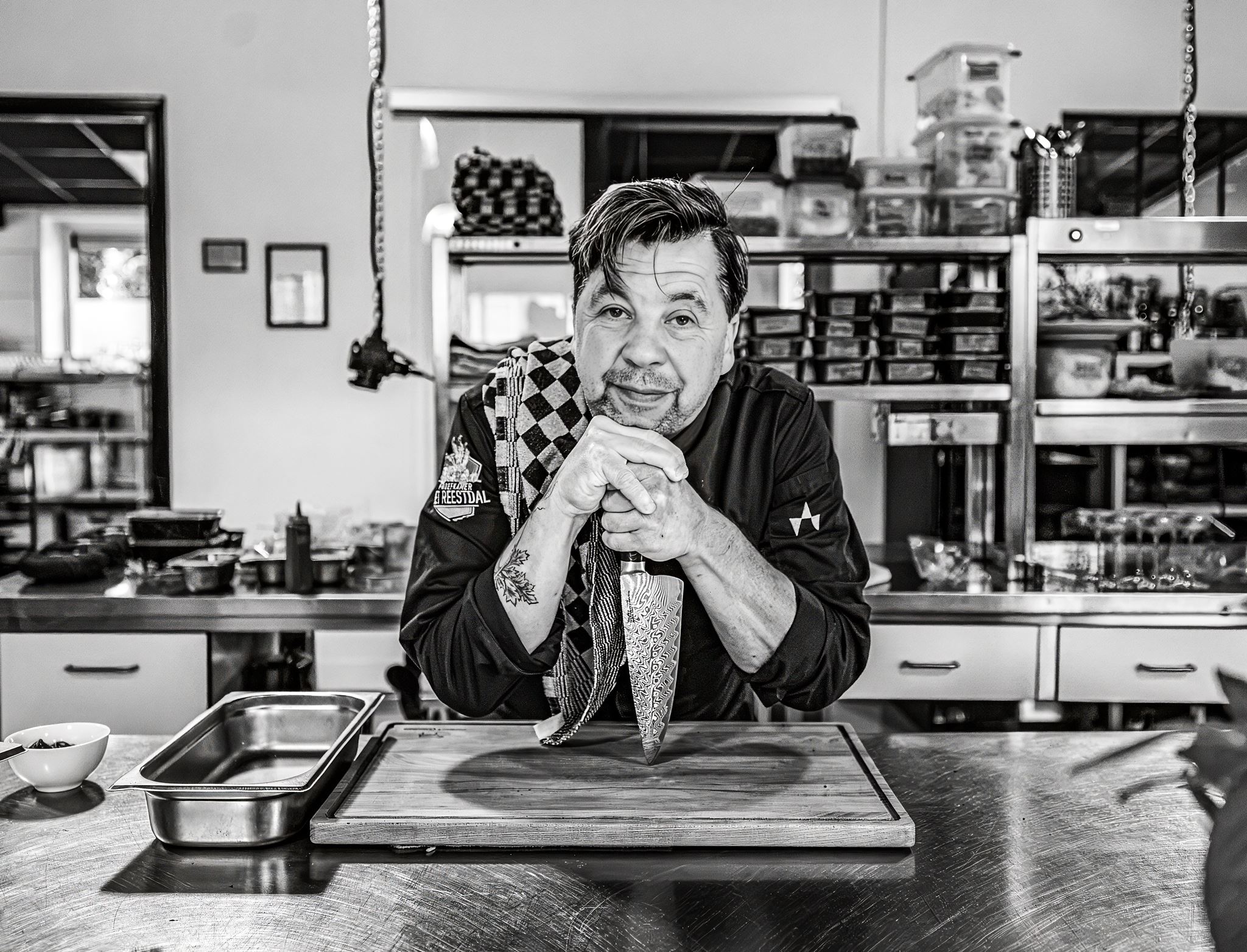 Chef leaning on a knife on a cutting board in a kitchen with a towel over his shoulder.