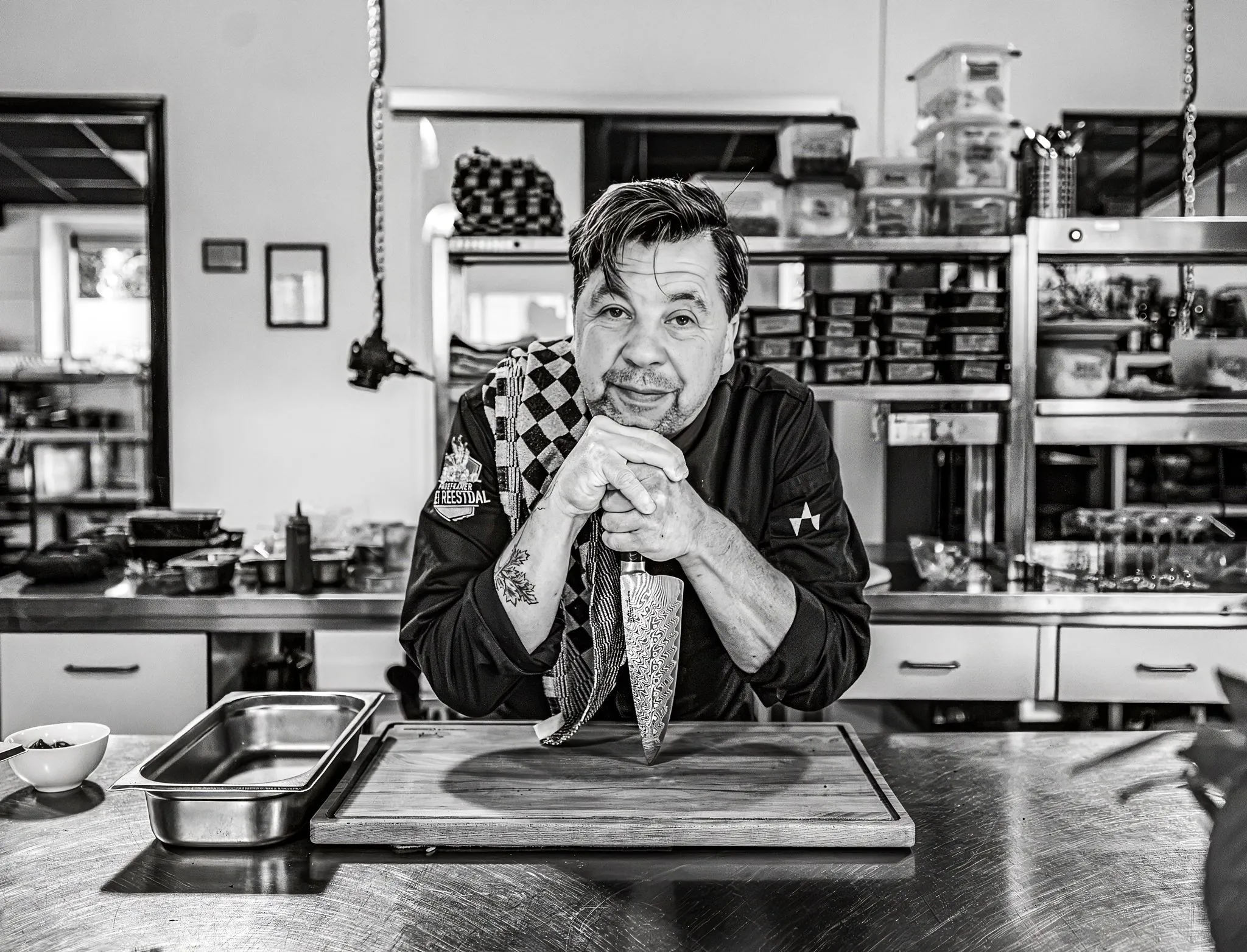 Chef leaning on a knife on a cutting board in a kitchen with a towel over his shoulder.