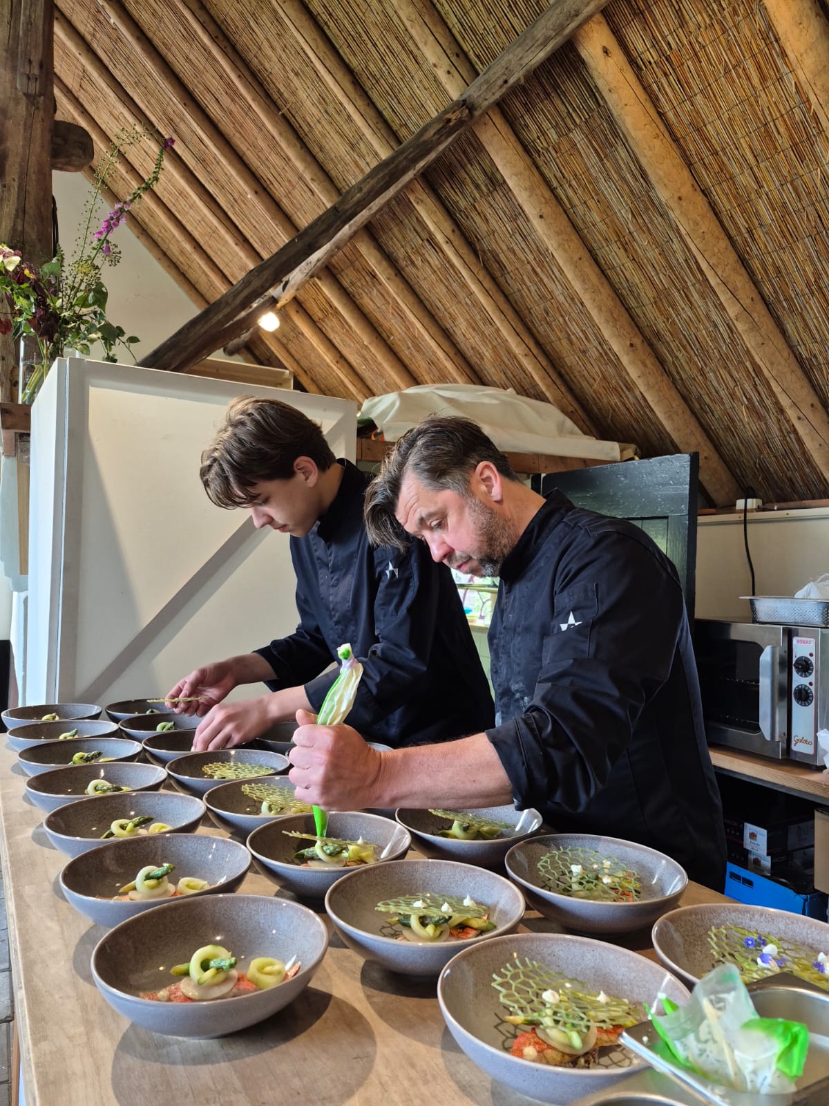 Two chefs in black uniforms plating gourmet dishes in gray bowls on a wooden table under a rustic ceiling.