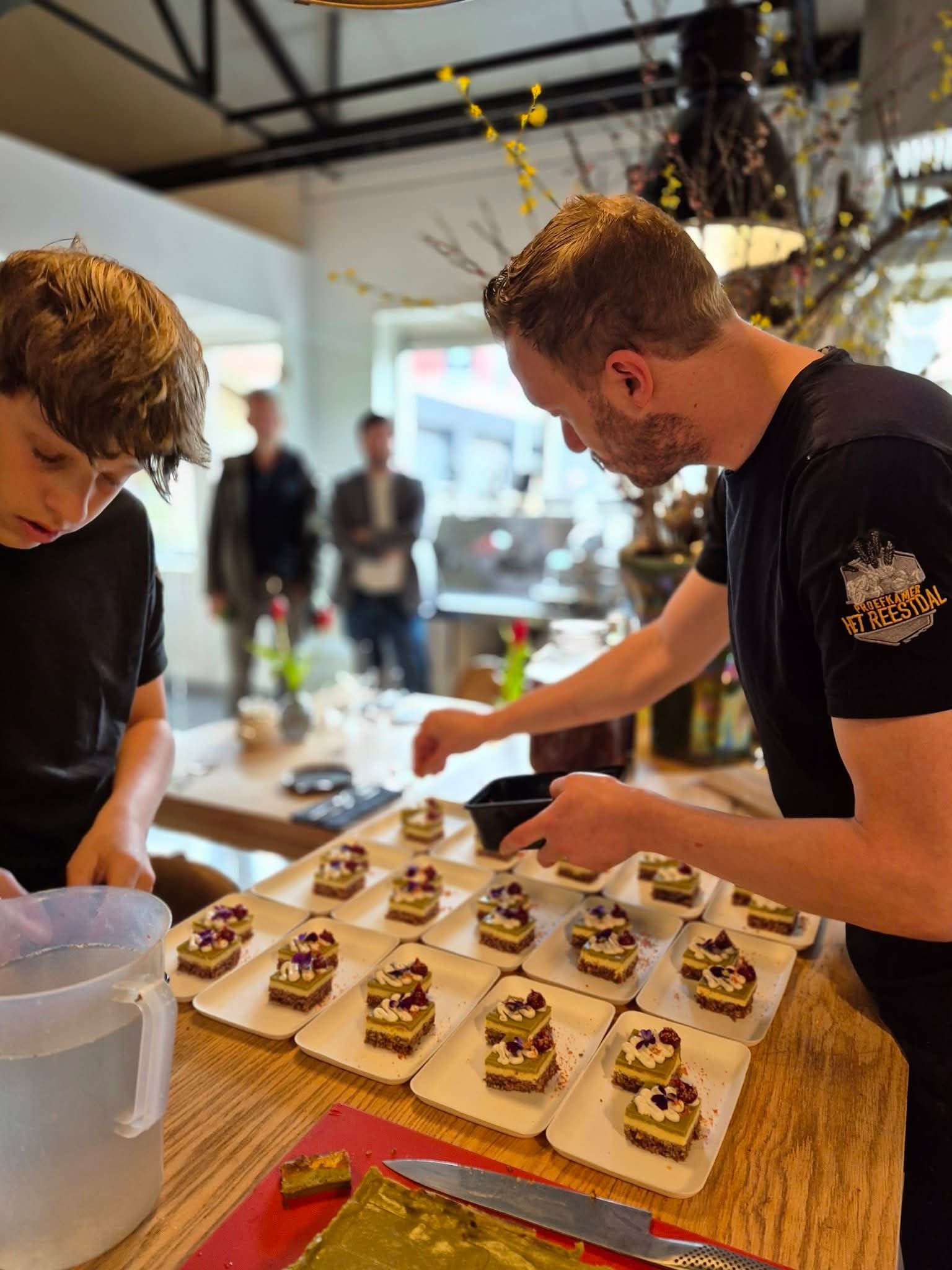 Two men plating square desserts with cream and flower decorations on individual plates.