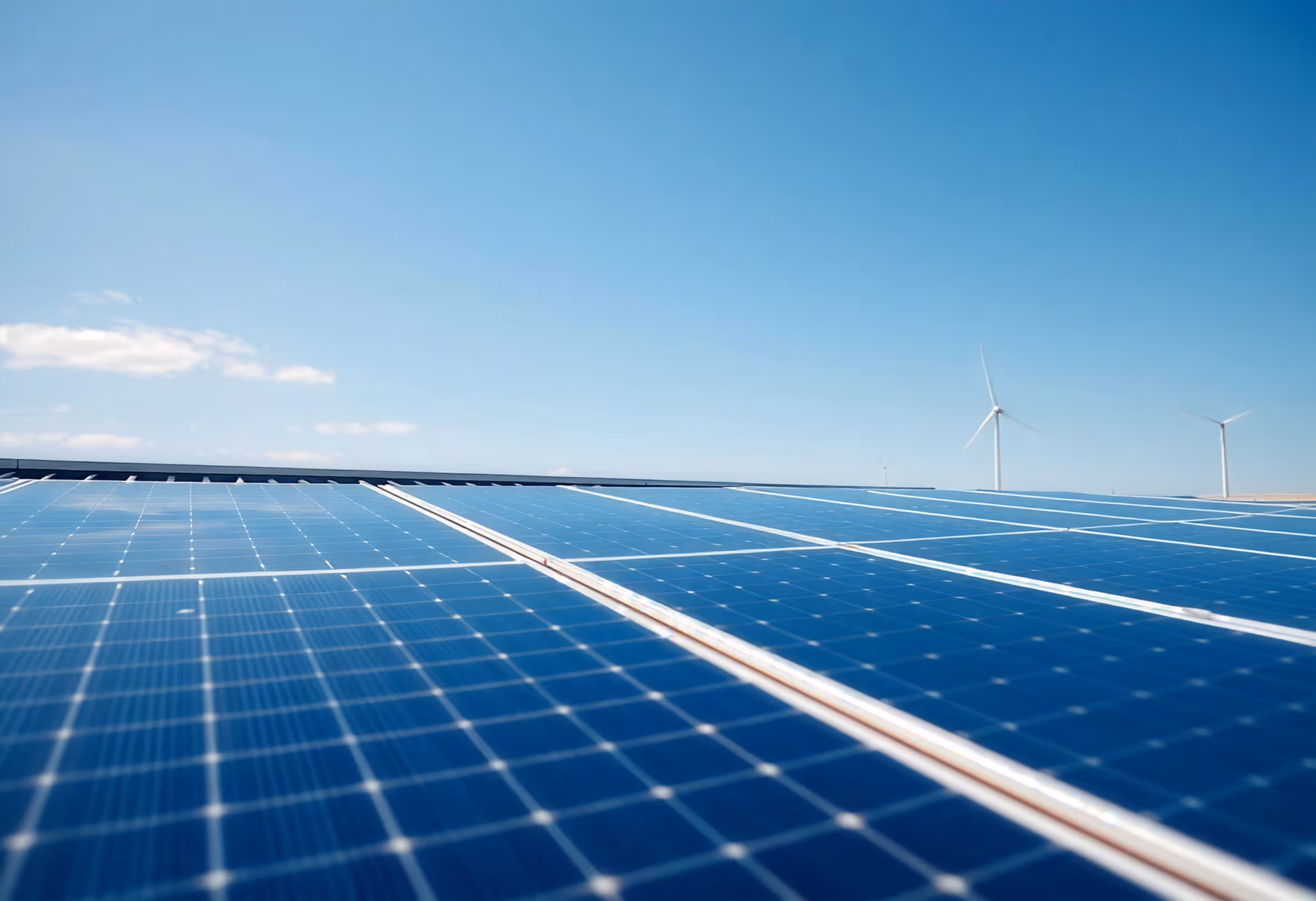 Close-up of solar panels with wind turbines in background under clear blue sky.