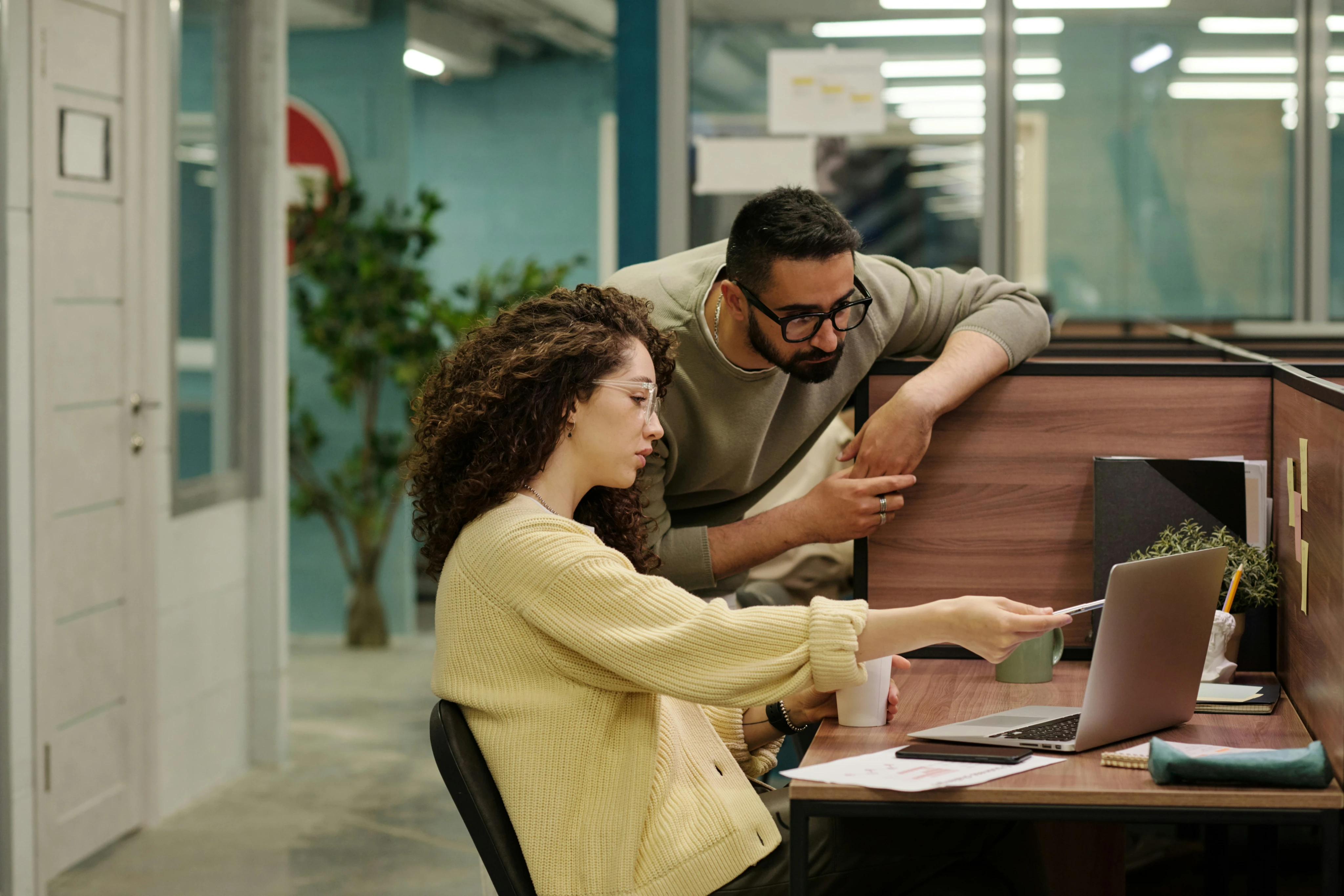 Two coworkers discussing work on a laptop in a modern office cubicle.