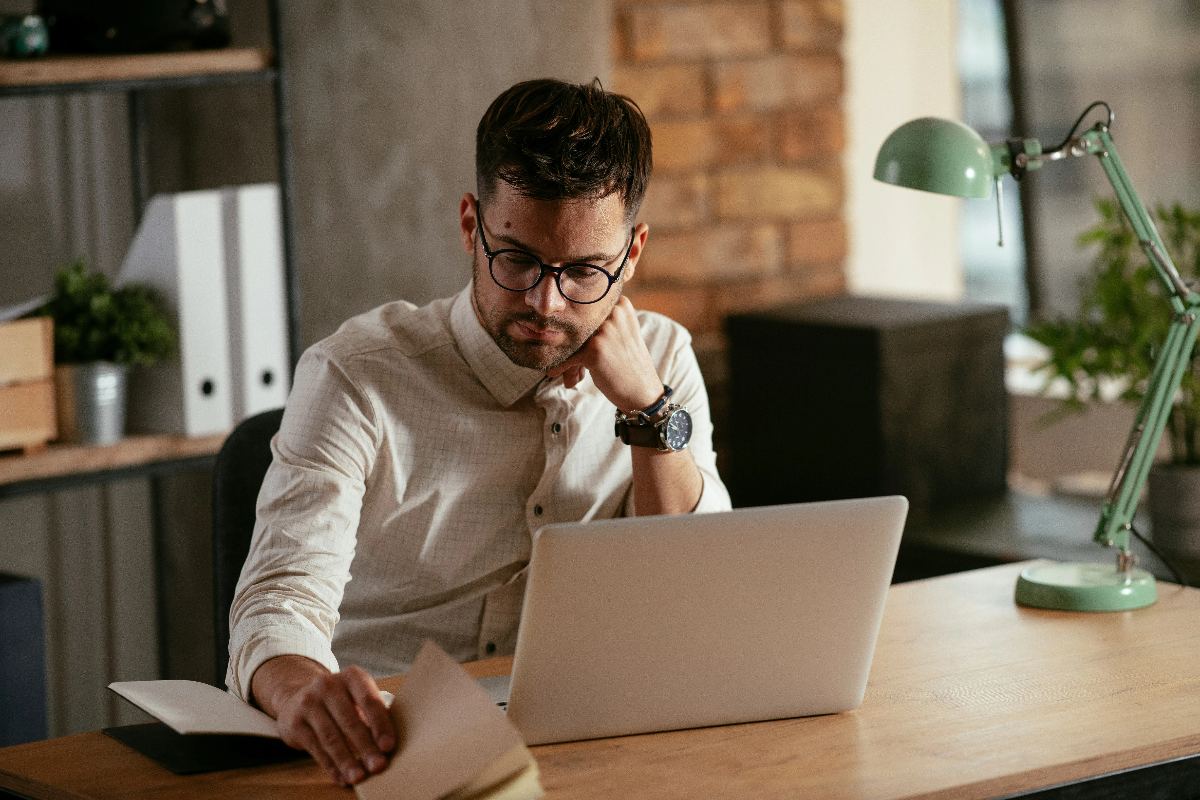 Man wearing glasses sits at desk with laptop, looking at papers in an office setting with lamp and plants.