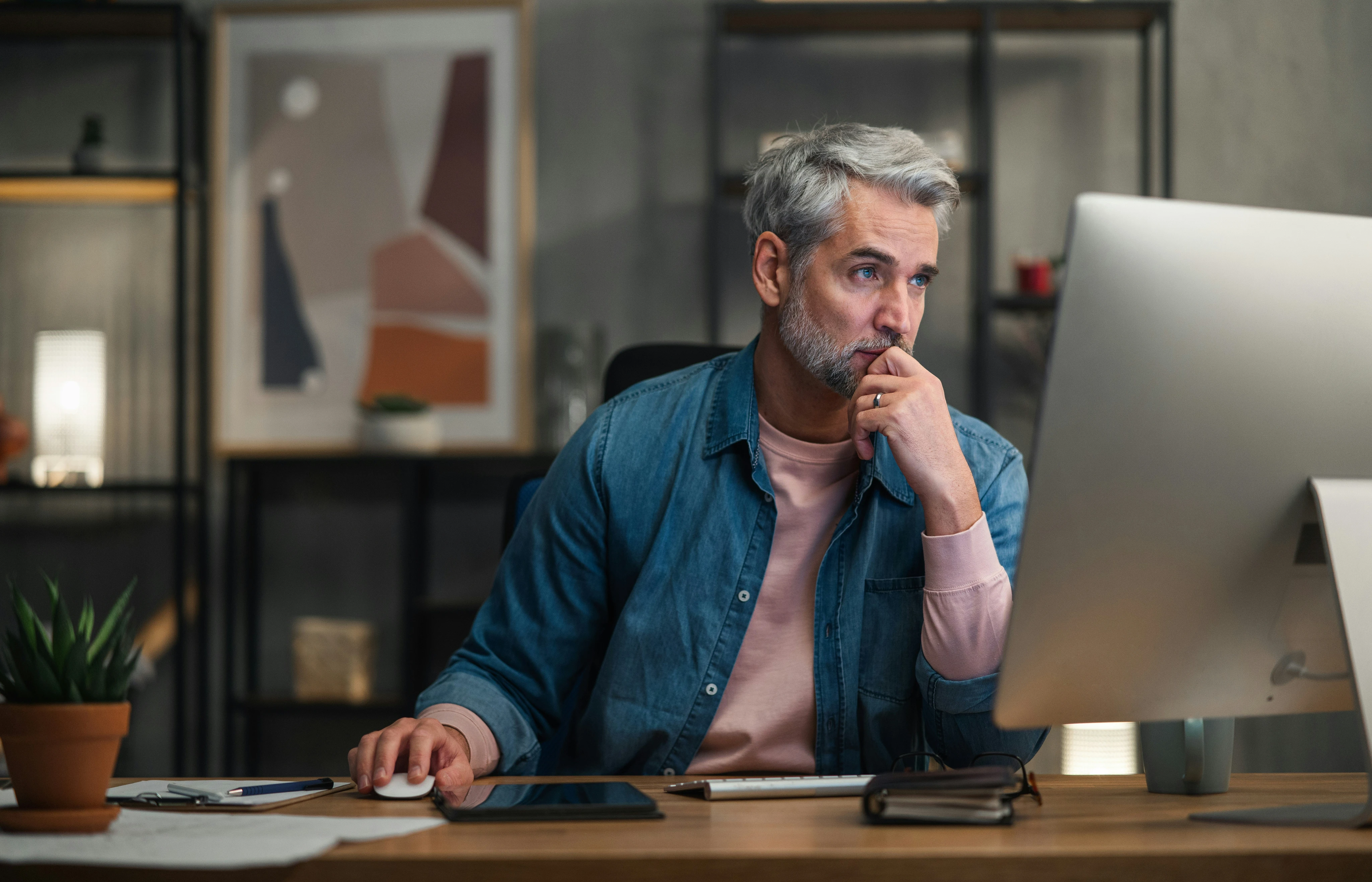 Thoughtful man with gray hair working on a desktop computer at a desk with office items.