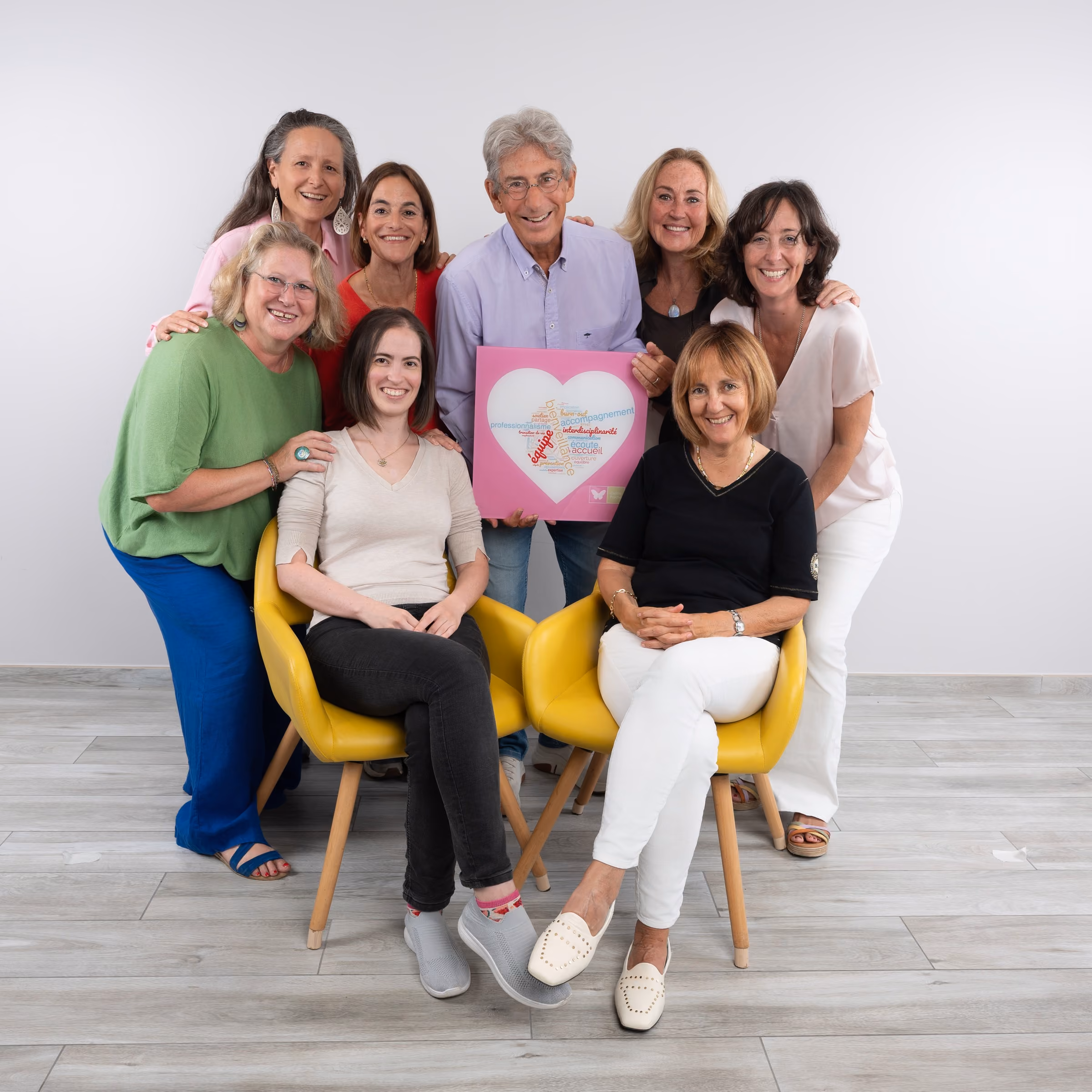 Sept personnes souriantes posent devant un mur blanc, deux femmes sont assises sur des fauteuils jaunes au premier plan, un homme et quatre femmes debout derrière tenant un panneau rose avec un cœur blanc contenant des mots colorés.
