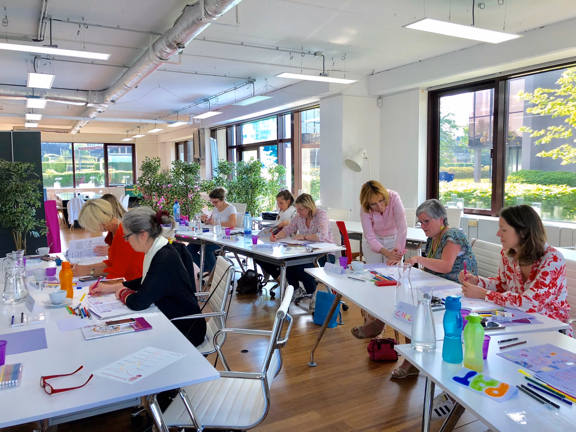 Salle lumineuse avec plusieurs femmes assises autour de tables blanches, participant à un atelier créatif ou de travail, en train d'écrire ou dessiner, avec des plantes vertes et de grandes fenêtres laissant entrer la lumière naturelle.