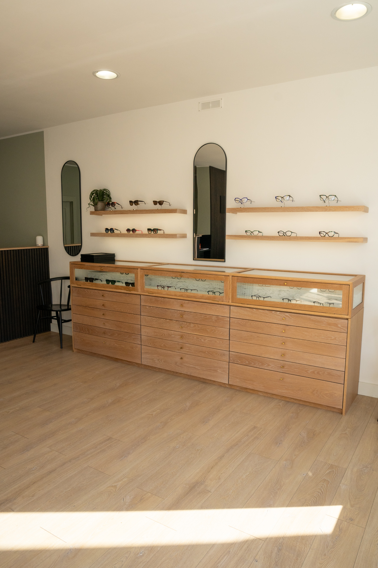 Wooden display cabinet with eyeglasses, two mirrors, shelves with more glasses, and a black chair in a bright room.