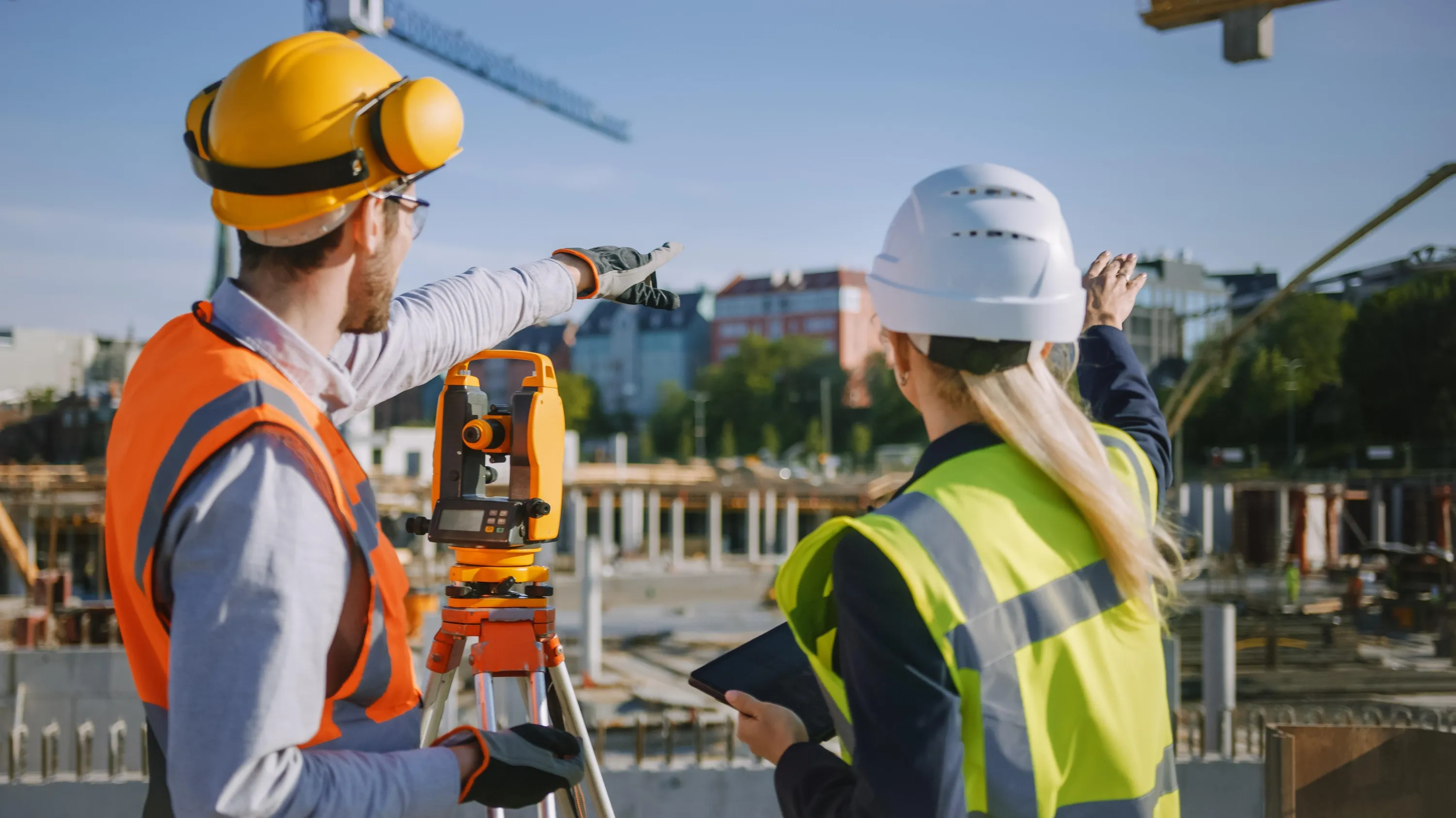 Kaksi rakennustyöntekijää takaapäin rakennustyömaalla aurinkoisena päivänä. Vasemmalla mies keltaisessa kypärässä ja oranssissa huomioliivissä osoittaa eteenpäin. Oikealla nainen valkoisessa kypärässä ja keltaisessa huomioliivissä pitää tablettia ja osoittaa ylöspäin. Heidän välissään on oranssi ja musta takymetri kolmijalalla. Taustalla näkyy nosturi, rakennuksia ja sininen taivas.