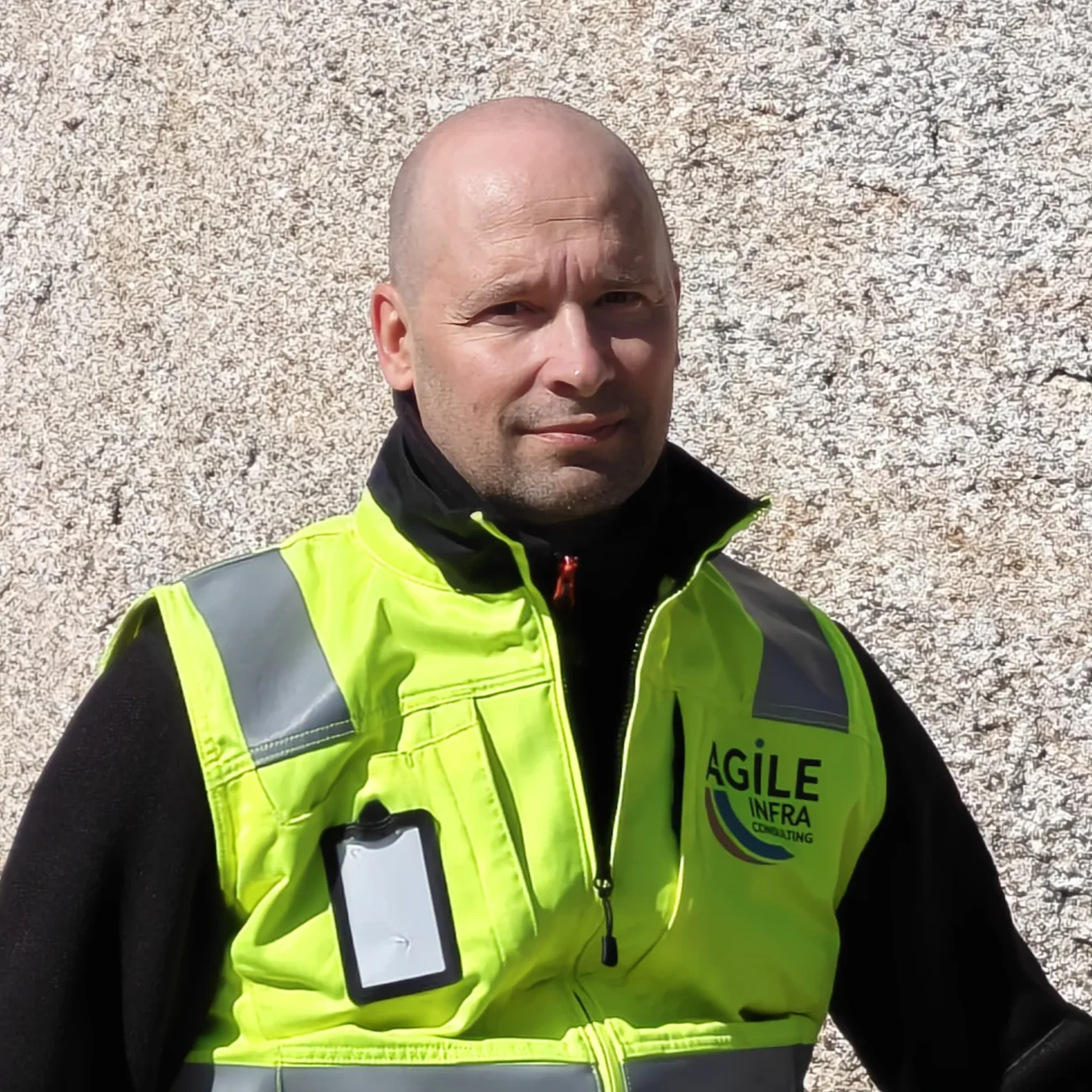 Lähikuva kaljusta, partasänkisestä miehestä, joka katsoo suoraan kameraan. Hänellä on yllään musta paita ja kirkkaan keltainen huomioliivi, jossa on harmaat heijastinnauhat. Liivin vasemmassa rintataskussa on tyhjä nimikylttipidike ja oikeassa rinnassa "AGILE INFRA CONSULTING" -logo. Taustalla on vaalea, karhea seinä.